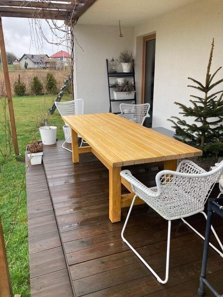 Zadaszony taras z drewnianą podłogą, na której stoi jasny, drewniany stół z czterema białymi krzesłami z plecionki, w tle widoczny ogród i dom.