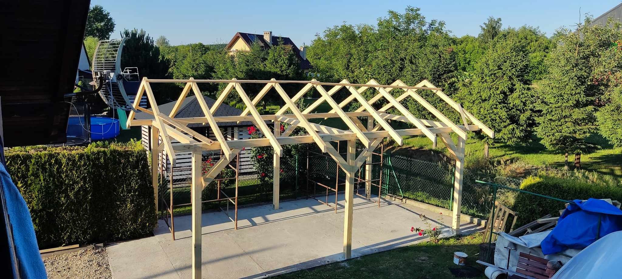 Konstrukcja drewniana zadaszenia w trakcie budowy, widoczne elementy szkieletu, słupy nośne, w tle dom i zieleń ogrodu.