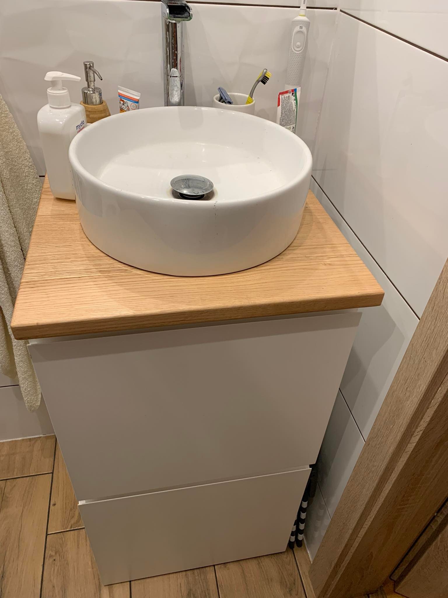 Biała, okrągła umywalka nablatowa na blacie z jasnego drewna, zamontowana na białej szafce łazienkowej z szufladami, widoczna w łazience z białymi płytkami.