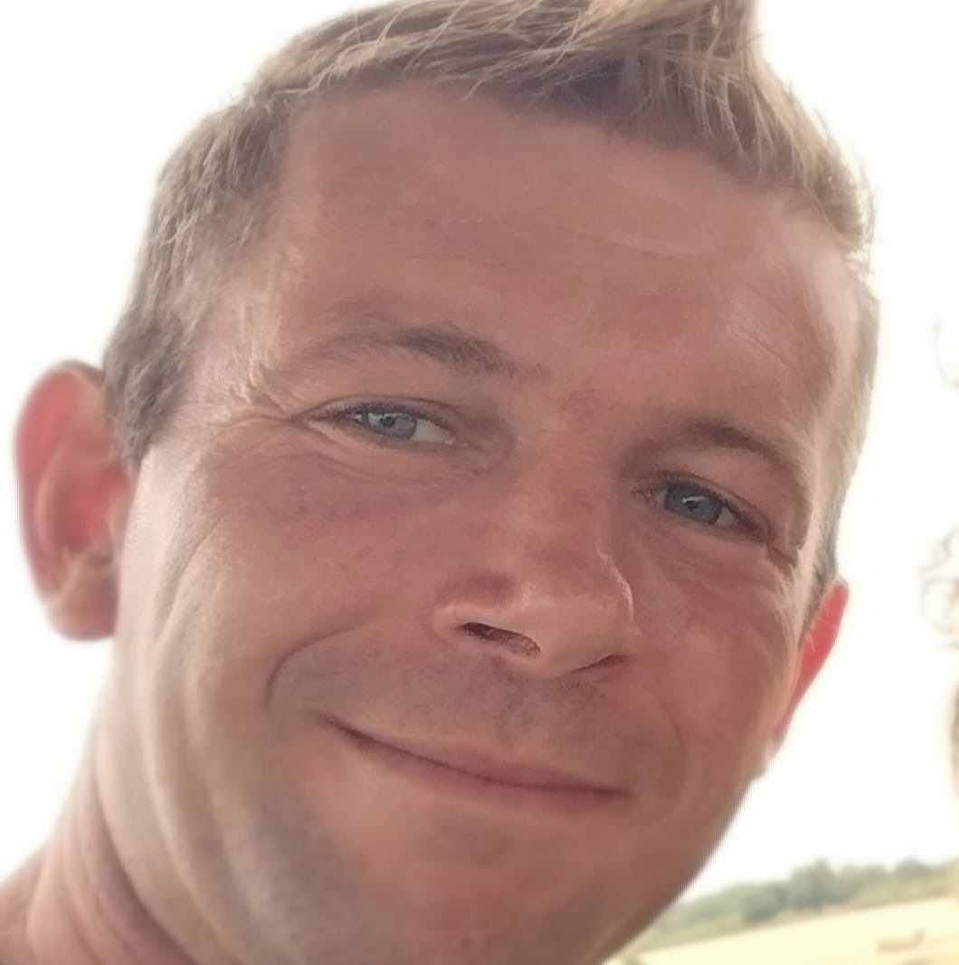 Portret uśmiechniętego mężczyzny z jasnymi włosami i niebieskimi oczami, z widocznymi zmarszczkami mimicznymi wokół oczu, na rozmytym tle krajobrazu.