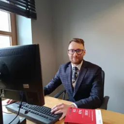 Uśmiechnięty mężczyzna w okularach i garniturze siedzi przy biurku z komputerem i kodeksem karnym na wierzchu.