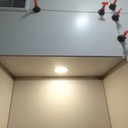 Świeżo ułożone, jasnoszare płytki na ścianie z wnęką oświetloną punktowym światłem, widoczne pomarańczowe klipsy poziomujące i sznurki między nimi.