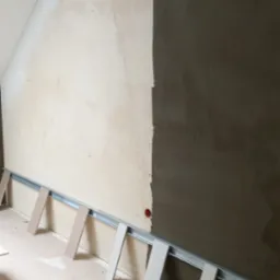 Ściana w trakcie szpachlowania, widoczne profile metalowe, płyty gipsowe, poziomica, wiertarka, laser krzyżowy i akcesoria glazurnicze na podłodze.