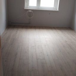 KONDIX KONRAD KOZUBSKI - Jasne panele podłogowe ułożone w pokoju z białymi ścianami i oknem. Widoczna listwa przypodłogowa i wystające przewody.  Podłoga gotowa.