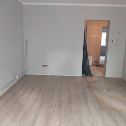 KONDIX KONRAD KOZUBSKI - Jasne panele podłogowe w szarym pokoju po remoncie. Widoczne rury grzewcze przy ścianie, niezabezpieczone gniazdka i przejście do kolejnego pomieszczenia.