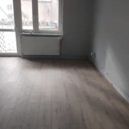 Ułożone panele podłogowe w szarym odcieniu w pokoju z oknem i grzejnikiem. Część paneli jeszcze nieułożona. Widoczne listwy przypodłogowe.