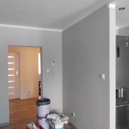 KONDIX KONRAD KOZUBSKI - Szare ściany z białymi listwami przypodłogowymi, widoczne narzędzia malarskie i wiadro z farbą. W tle widoczny fragment kuchni z lodówką.