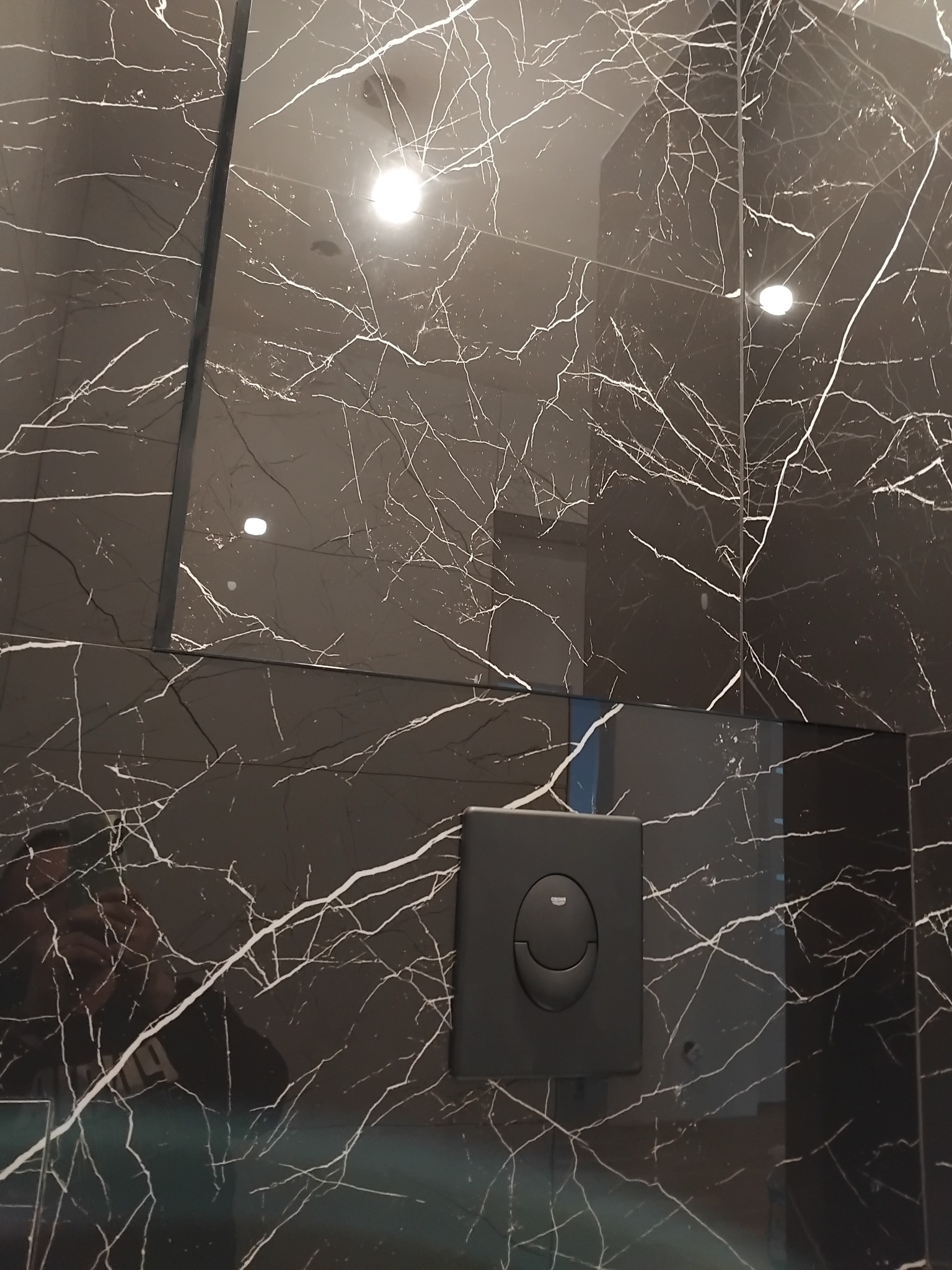 Czarne płytki marmurowe z białymi żyłami w łazience, z widocznym przyciskiem spłuczki Grohe. Nowoczesny design, wykończenie z połyskiem.