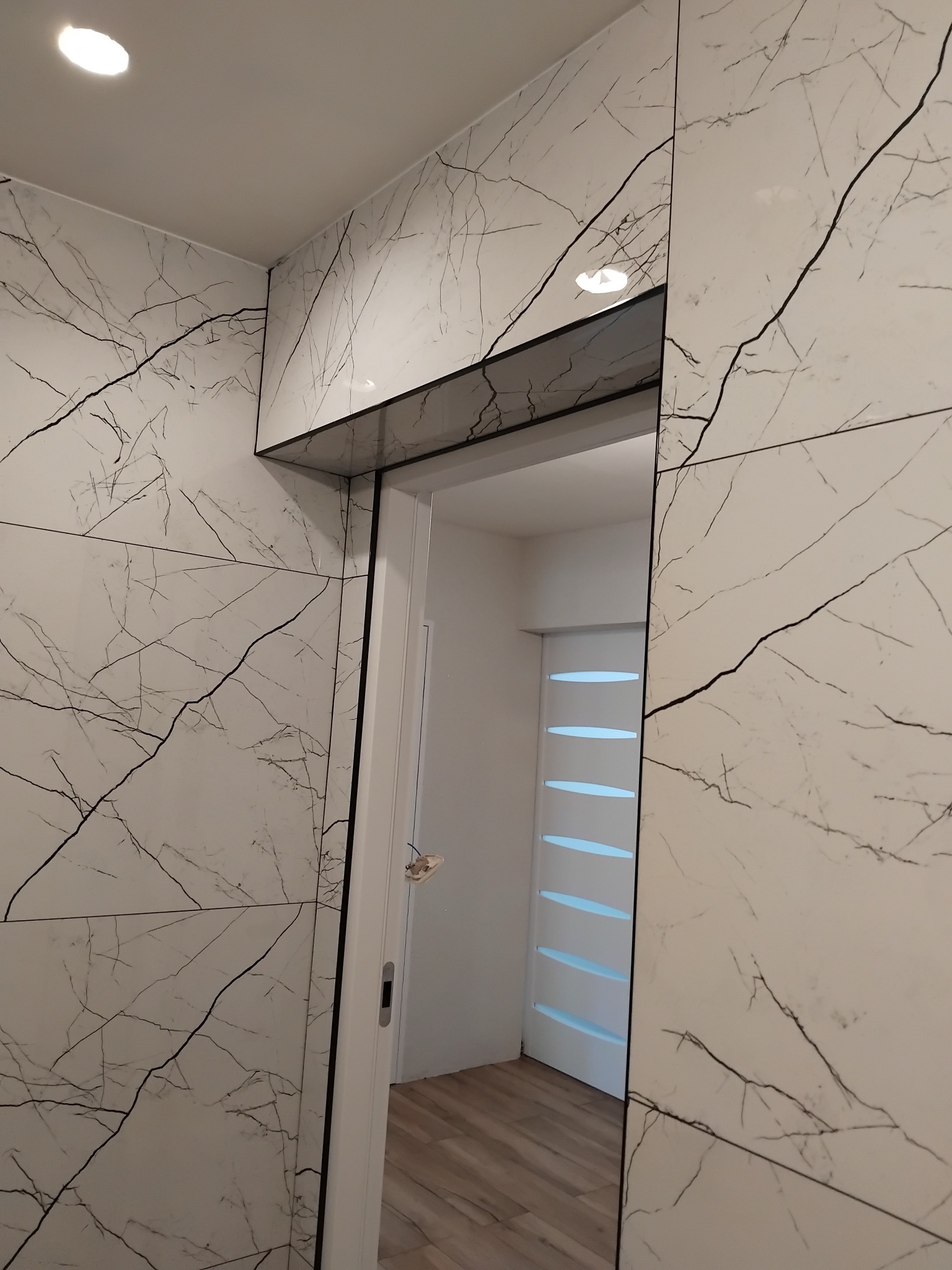Wykończenie łazienki z płytkami imitującymi marmur, widoczny fragment drzwi i oświetlenie sufitowe. Minimalistyczny design i precyzyjne ułożenie płytek.