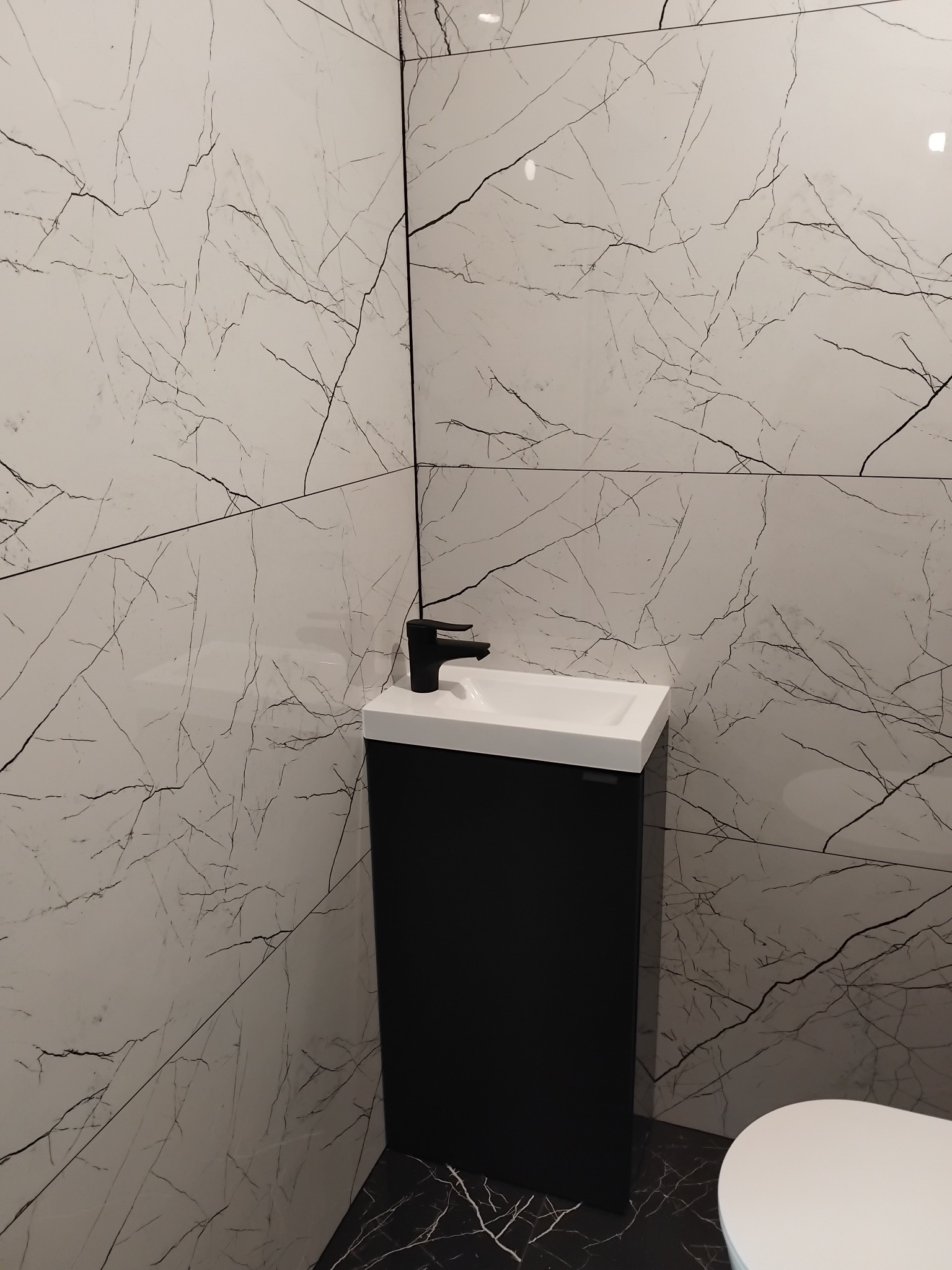 Nowoczesna łazienka z umywalką na szafce w kolorze czarnym, białą armaturą i płytkami imitującymi marmur na ścianach i podłodze. Minimalistyczny design.