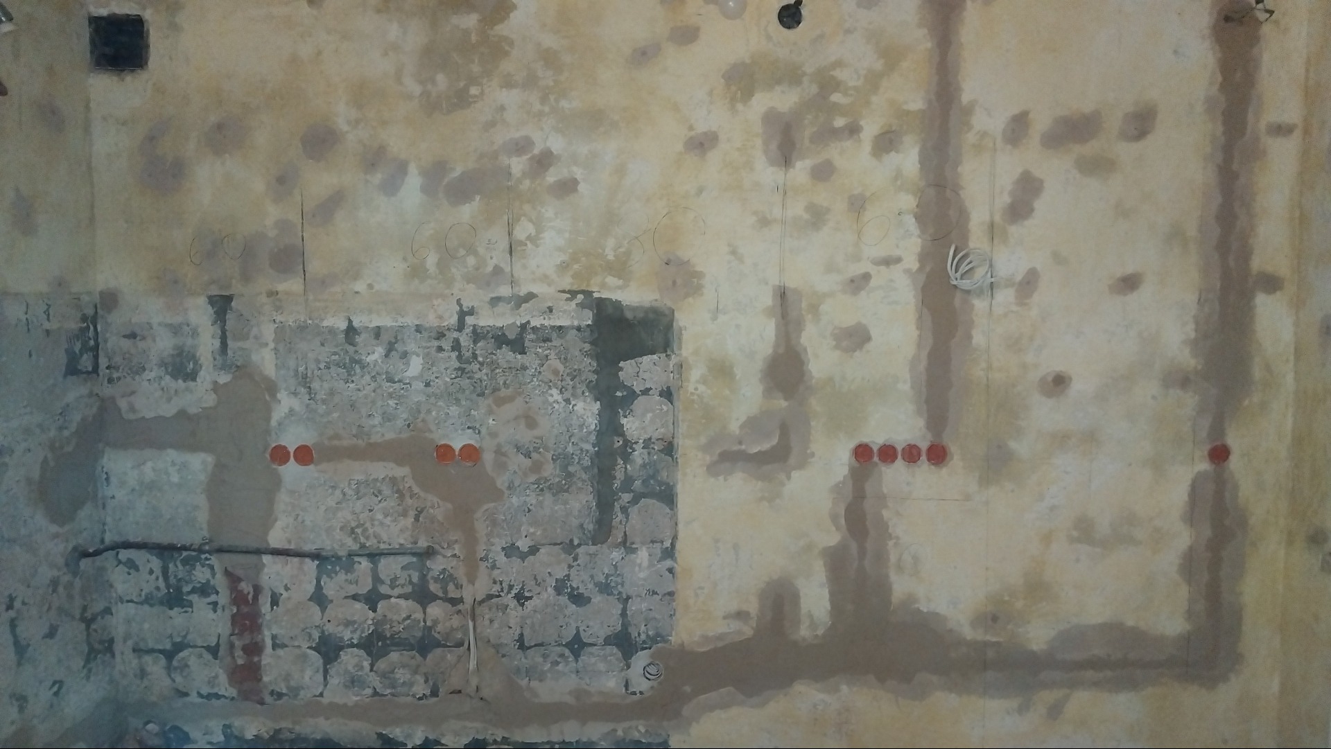 Ściana w trakcie przygotowań do prac wykończeniowych z widocznymi instalacjami elektrycznymi i fragmentami starej okładziny. Widoczne ślady po kuciu i bruzdach.