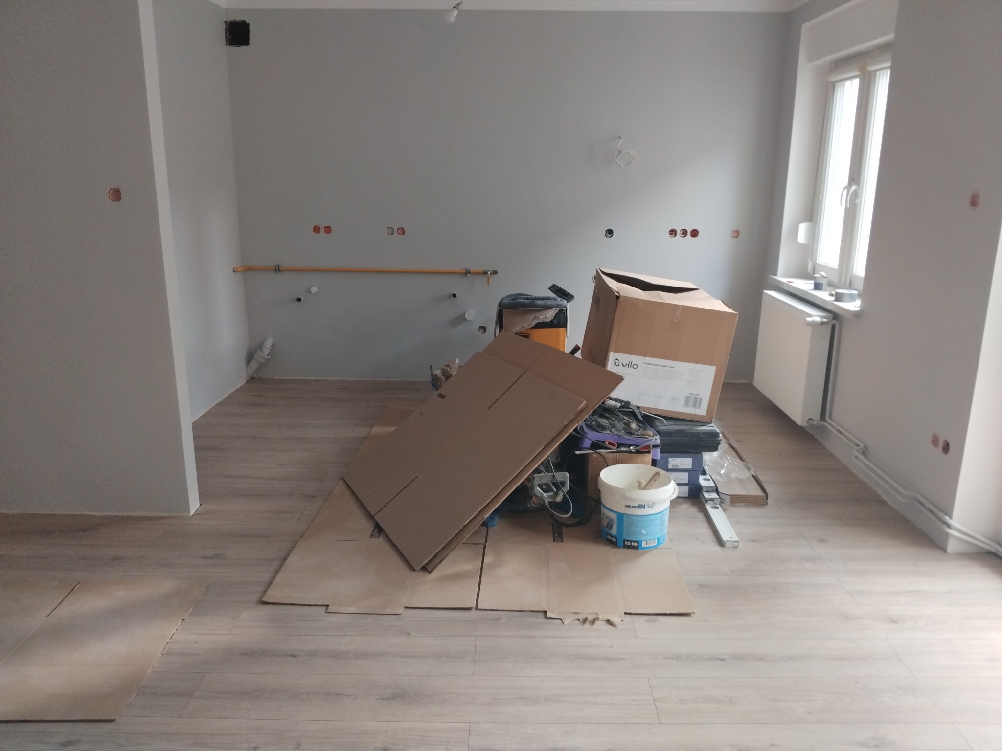 Przygotowania do remontu: zabezpieczona podłoga panelowa kartonem, widoczne narzędzia, puszki elektryczne i rury na ścianie w szarym pomieszczeniu.