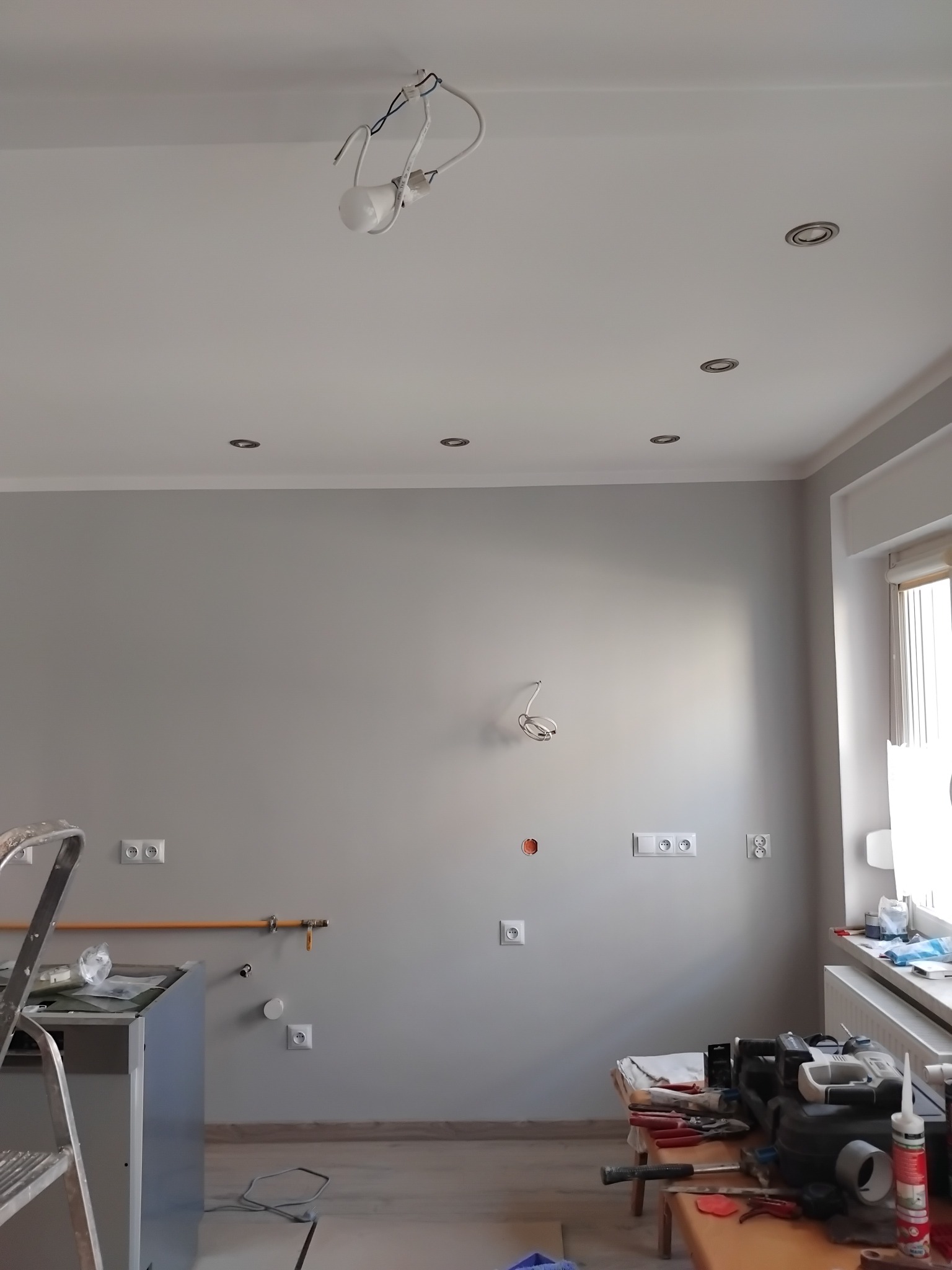 Szare wnętrze w trakcie prac wykończeniowych: widoczne instalacje elektryczne, rury, drabina i narzędzia na stole. Oświetlenie sufitowe częściowo zamontowane.