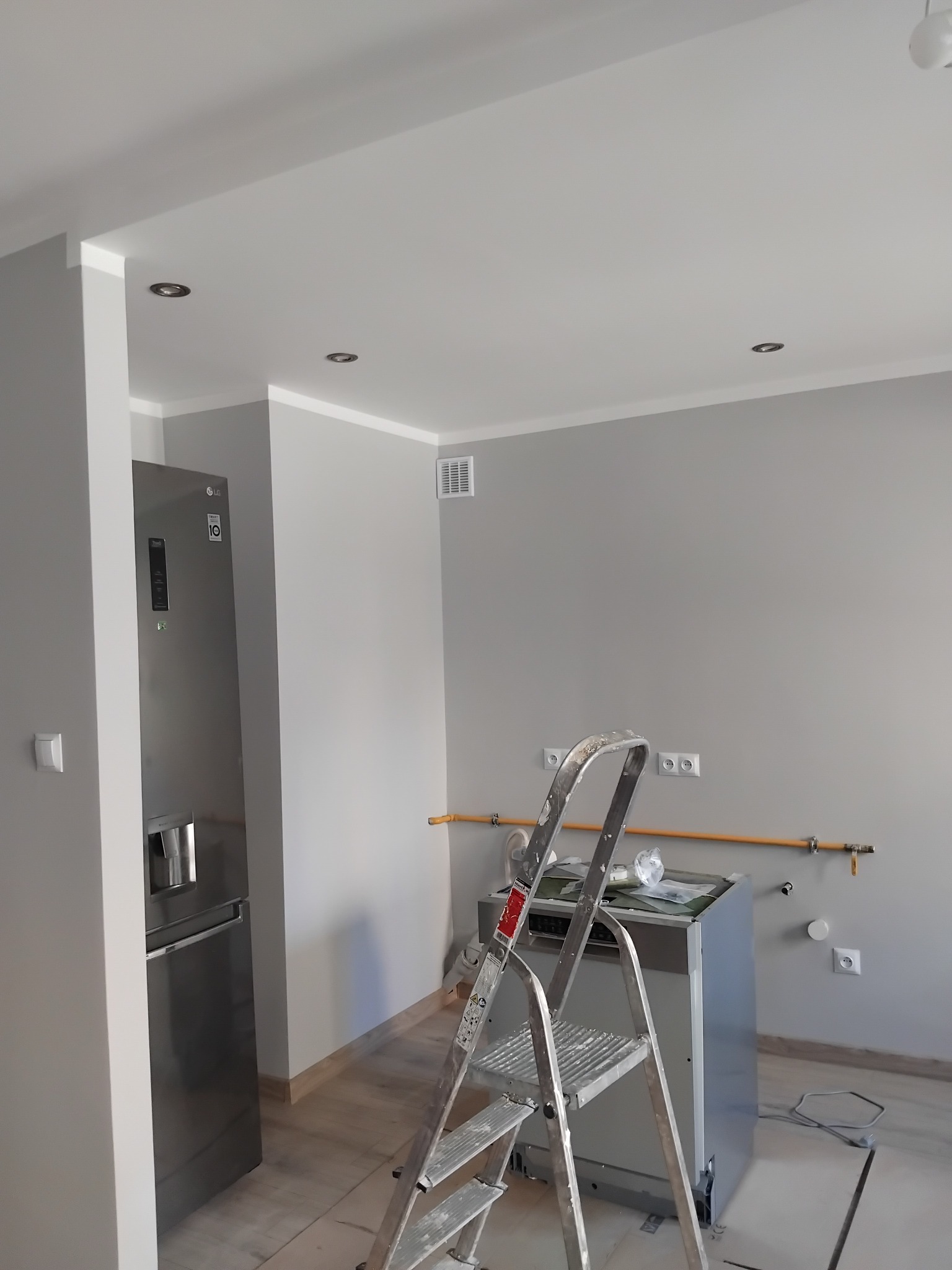Szaro-biała kuchnia w trakcie wykończenia: świeżo pomalowane ściany, sprzęt AGD, drabina malarska, widoczne instalacje, podwieszany sufit z oświetleniem.