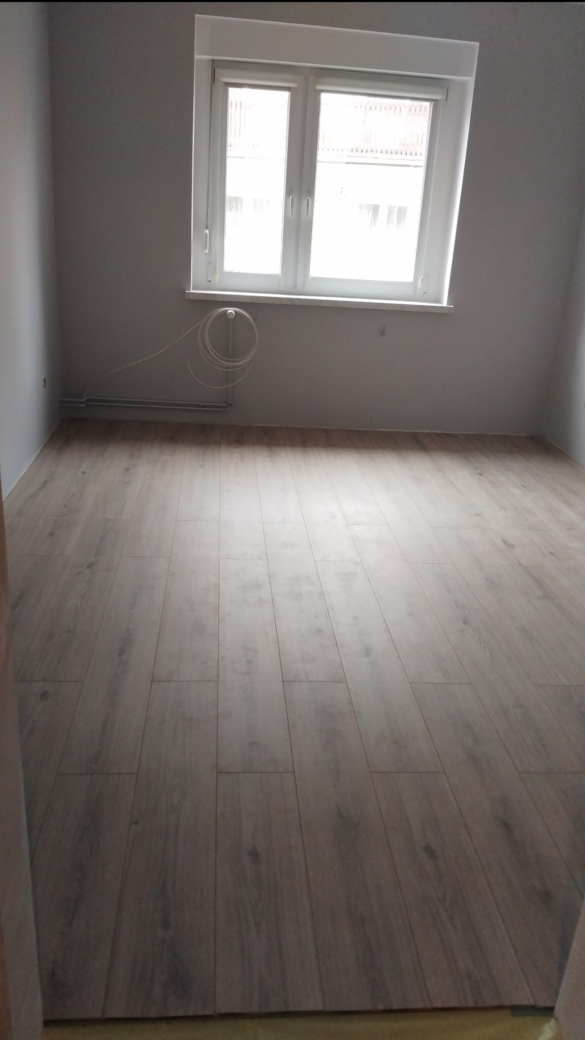 Jasne panele podłogowe ułożone w pokoju z białymi ścianami i oknem. Widoczna listwa przypodłogowa i wystające przewody.  Podłoga gotowa.