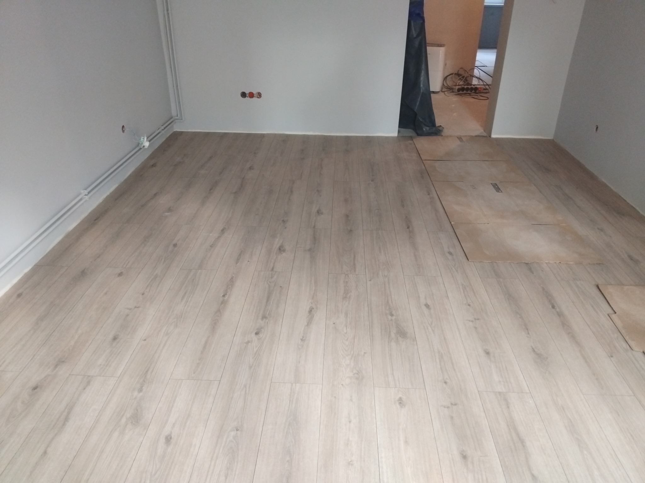 Jasne panele podłogowe w pokoju z szarymi ścianami. Część podłogi jeszcze nieukończona, widoczne płyty podkładowe. Minimalistyczne wnętrze w trakcie wykończenia.