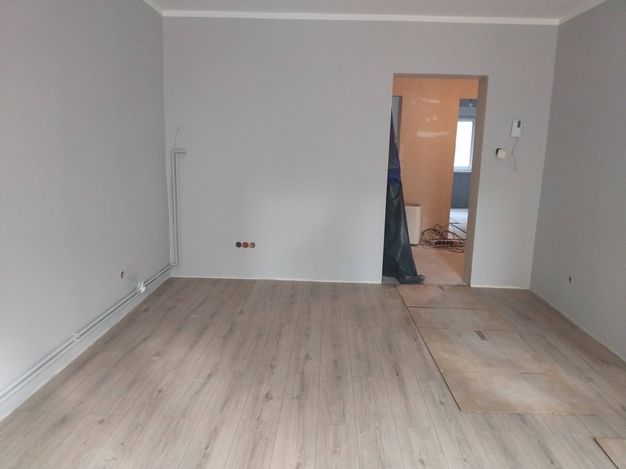 Jasne panele podłogowe w szarym pokoju po remoncie. Widoczne rury grzewcze przy ścianie, niezabezpieczone gniazdka i przejście do kolejnego pomieszczenia.