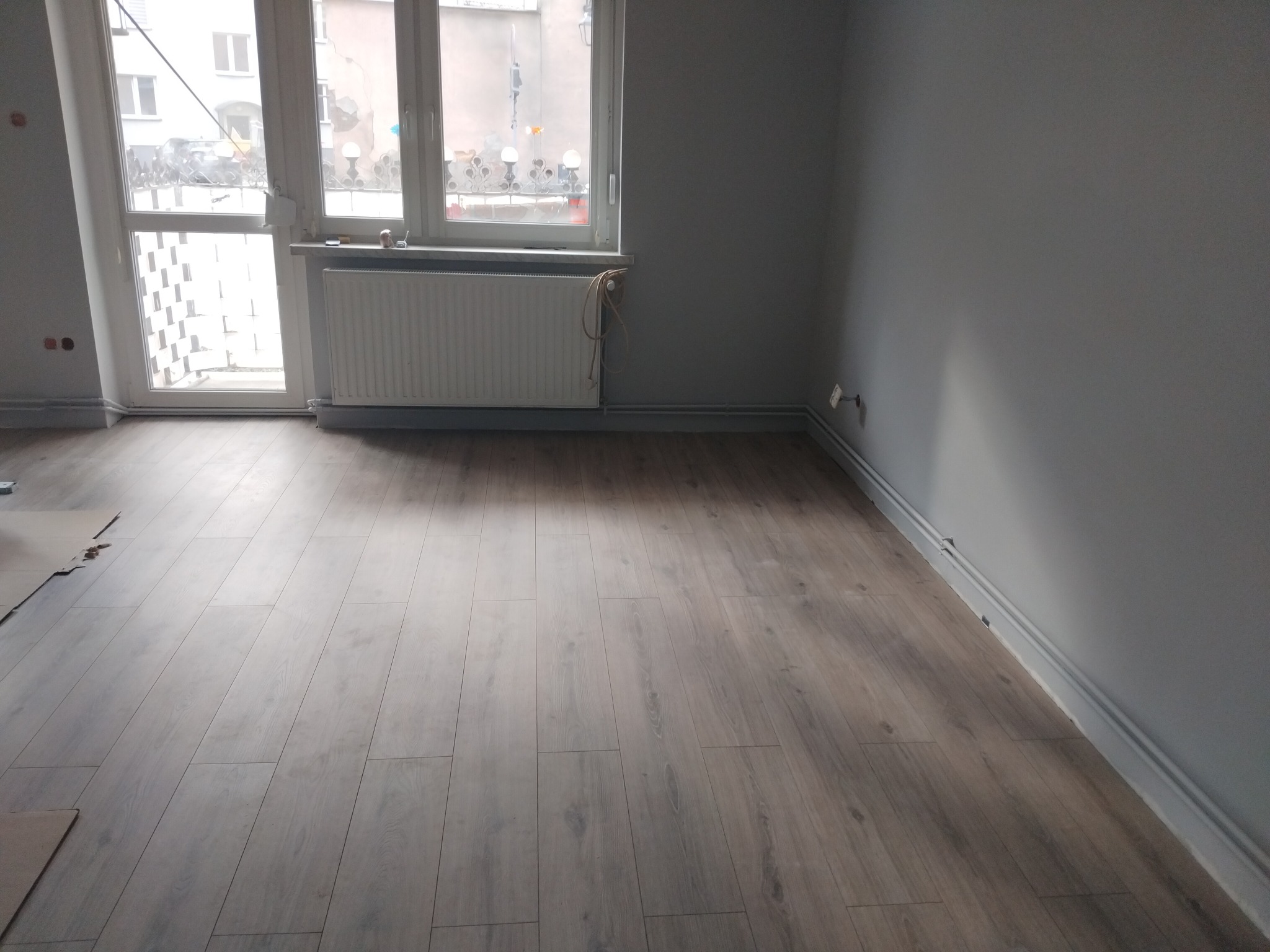 Ułożone panele podłogowe w szarym odcieniu w pokoju z oknem i grzejnikiem. Część paneli jeszcze nieułożona. Widoczne listwy przypodłogowe.