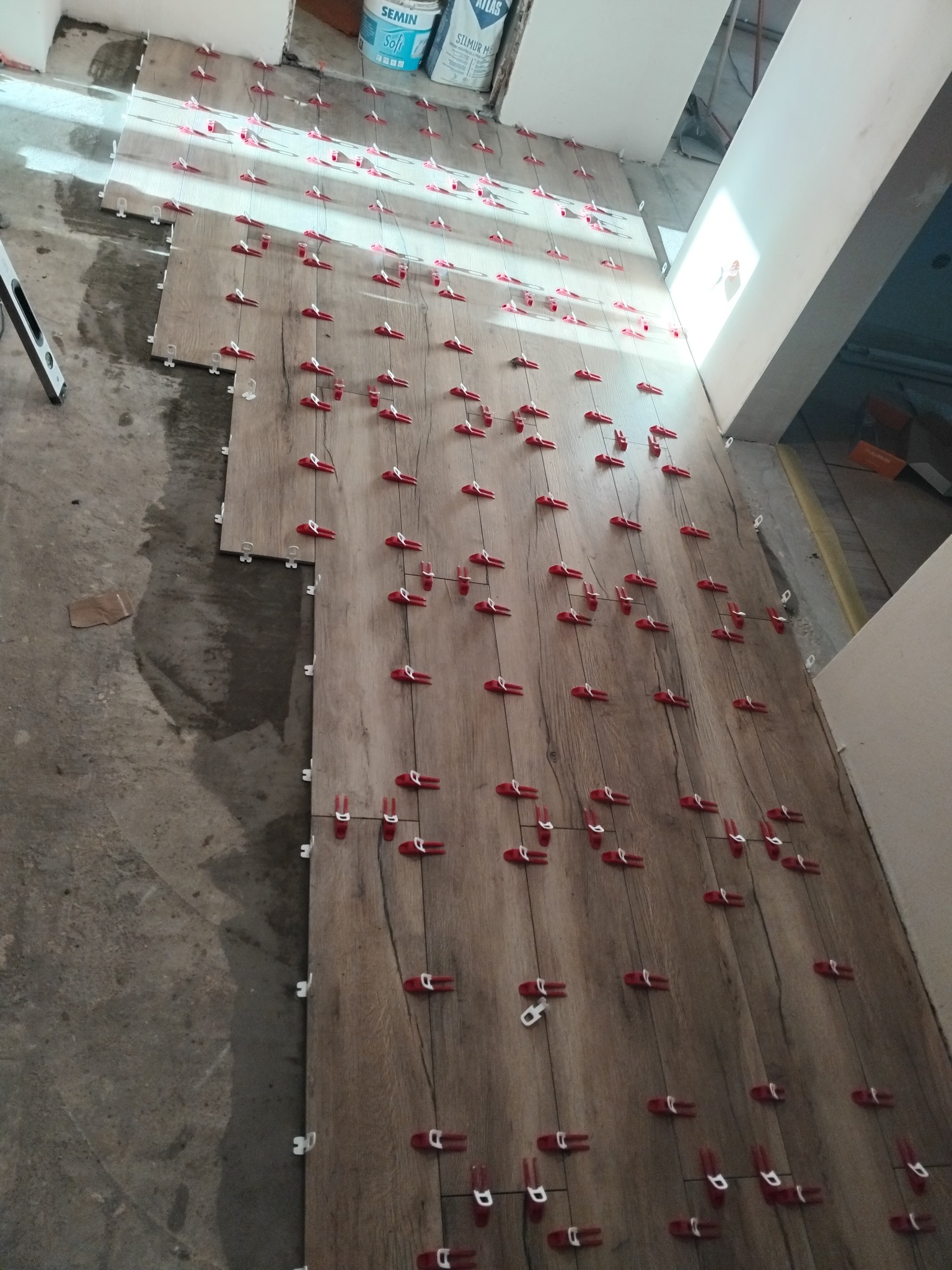 Układanie płytek imitujących drewno z użyciem czerwonych klipsów poziomujących. Częściowo ułożona podłoga, widoczne worki z zaprawą i poziomica.