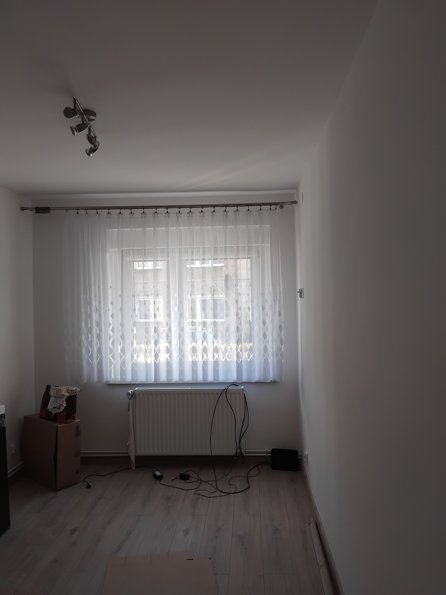 Jasne, puste wnętrze pokoju z panelami podłogowymi i oknem z białymi, wzorzystymi firanami. Ściany świeżo pomalowane na biało. Widoczny grzejnik i lampa sufitowa.