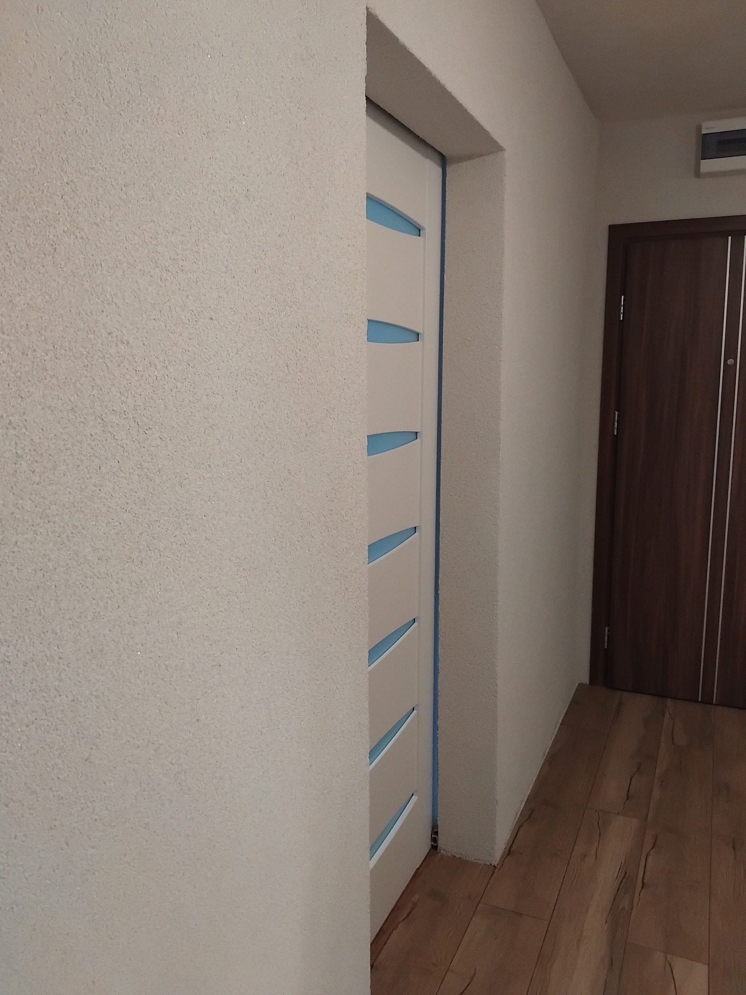 Białe drzwi wewnętrzne z przeszkleniami w kolorze błękitnym, widoczne na tle ściany o fakturze baranka i drewnopodobnej podłogi w mieszkaniu.