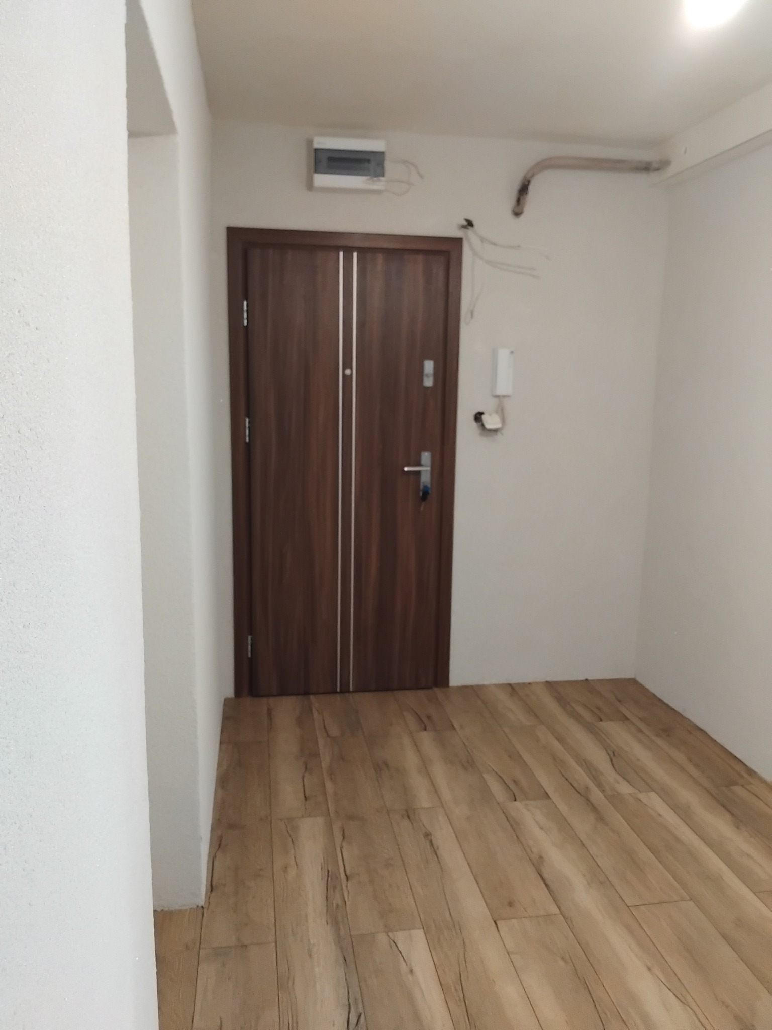 Wykończone wnętrze przedpokoju: brązowe drzwi z srebrnymi paskami, panele podłogowe imitujące drewno, białe ściany. Widoczne elementy instalacji elektrycznej.