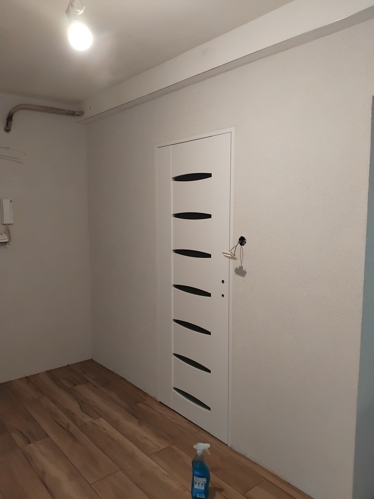Białe drzwi z poziomymi przeszkleniami w jasnym wnętrzu po remoncie. Na podłodze panele, na ścianie widoczne kable. Butelka płynu do czyszczenia na podłodze.