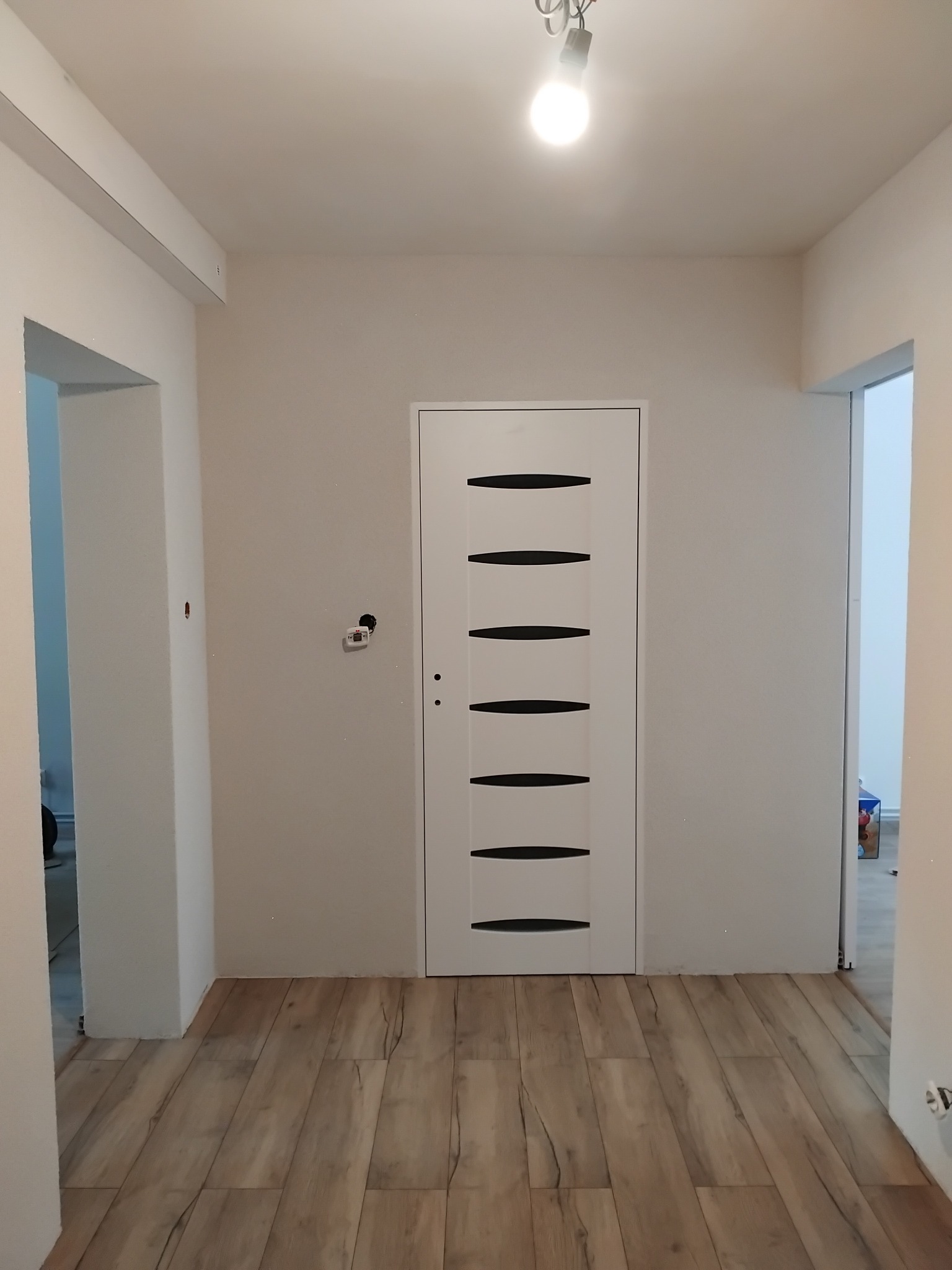 Wnętrze po remoncie: drewnopodobne panele, białe ściany i nowoczesne drzwi z czarnymi akcentami. Widoczne niedokończone detale wykończeniowe.