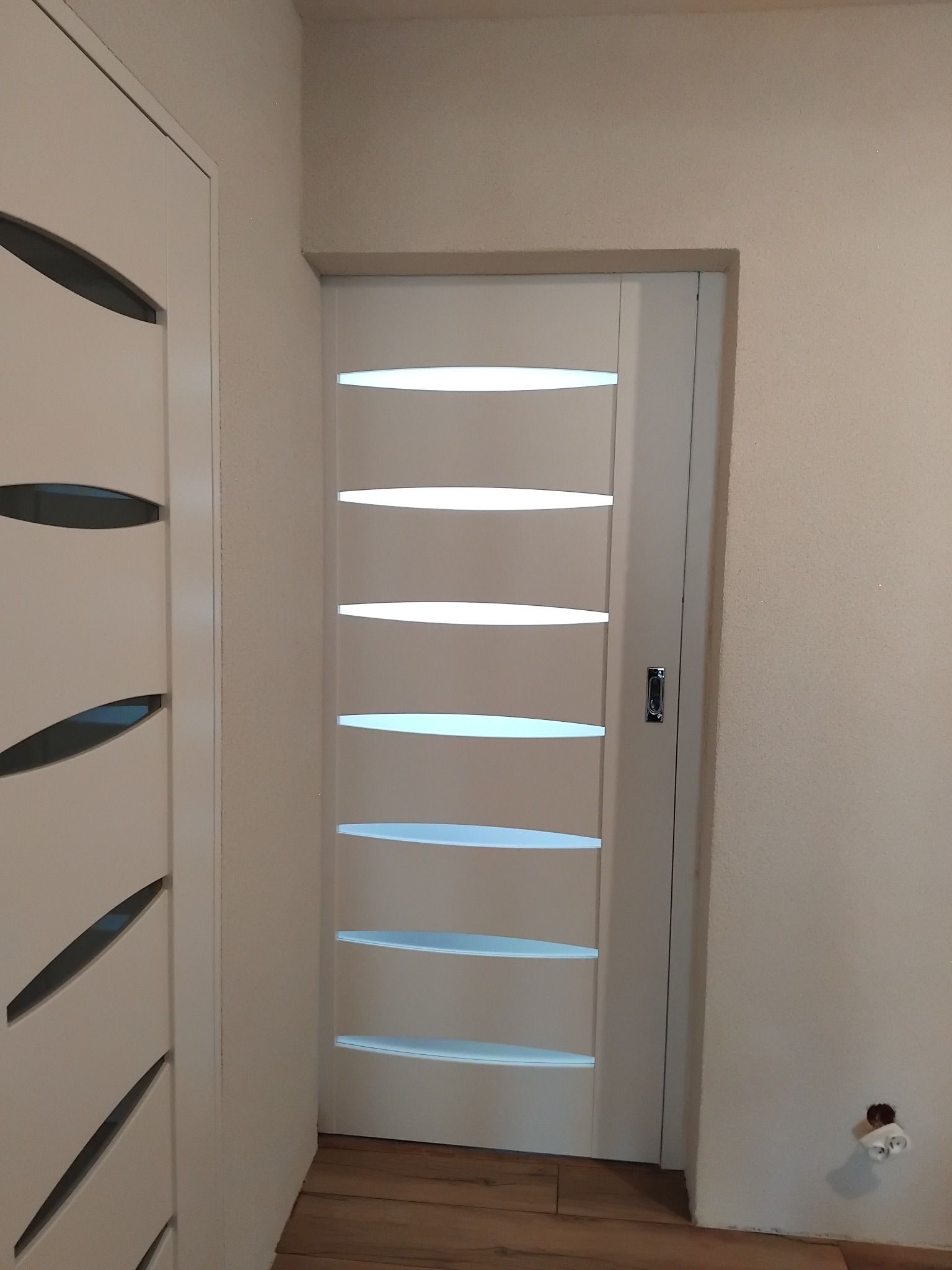 Białe, nowoczesne drzwi wewnętrzne z przeszkleniami w kształcie elips, zamontowane w jasnym pomieszczeniu. Widoczny fragment drugiej pary drzwi po lewej.