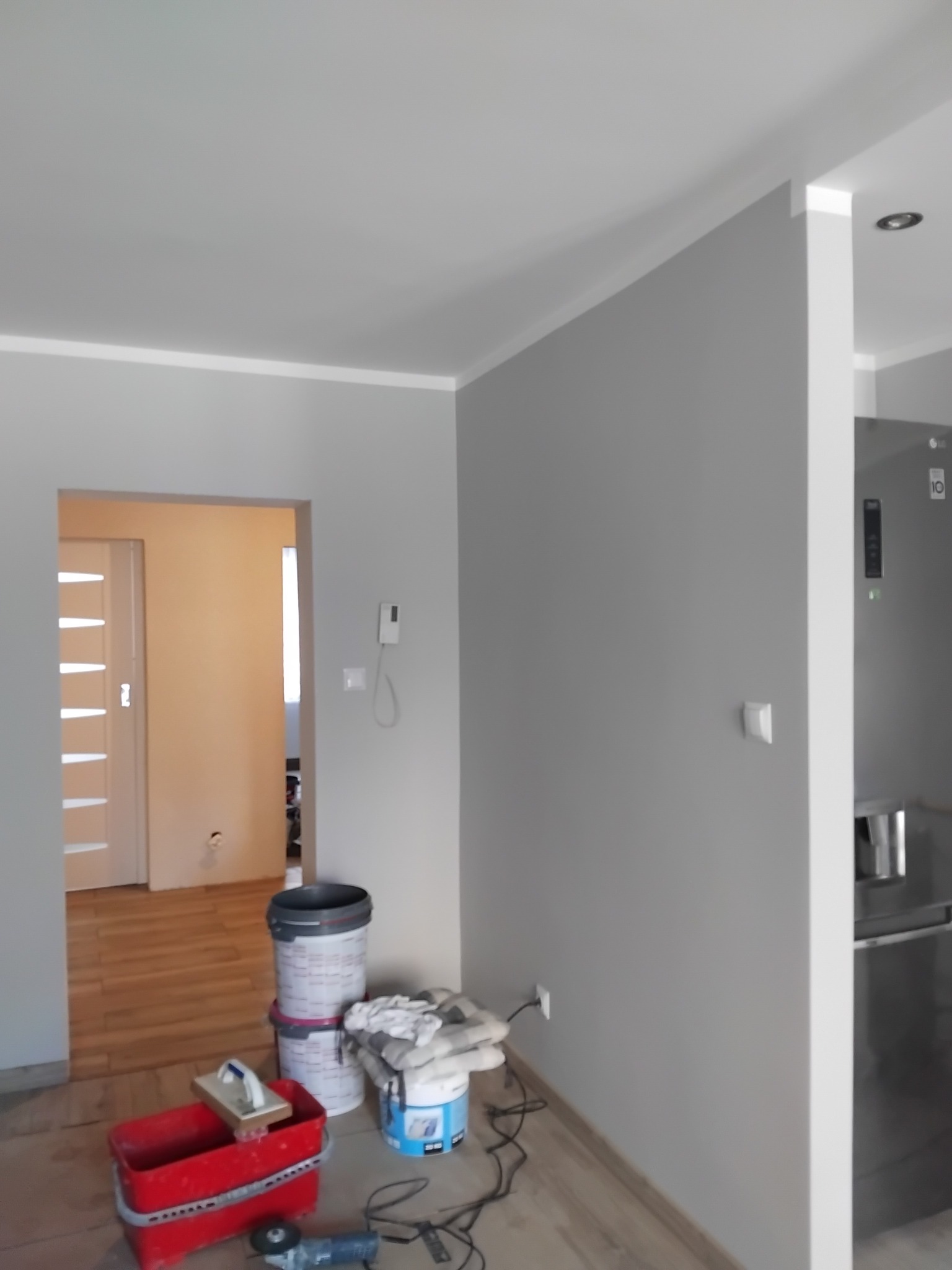 Szare ściany z białymi listwami przypodłogowymi, widoczne narzędzia malarskie i wiadro z farbą. W tle widoczny fragment kuchni z lodówką.