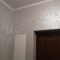 Kamil Stolat - Ściana wewnętrzna wykończona dekoracyjną, srebrzystą fakturą, widoczny fragment brązowych drzwi i biała skrzynka rewizyjna.