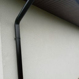 Kamil Stolat - Fragment jasnej elewacji budynku z widoczną czarną rynną spustową i brązowym podbitką dachową.