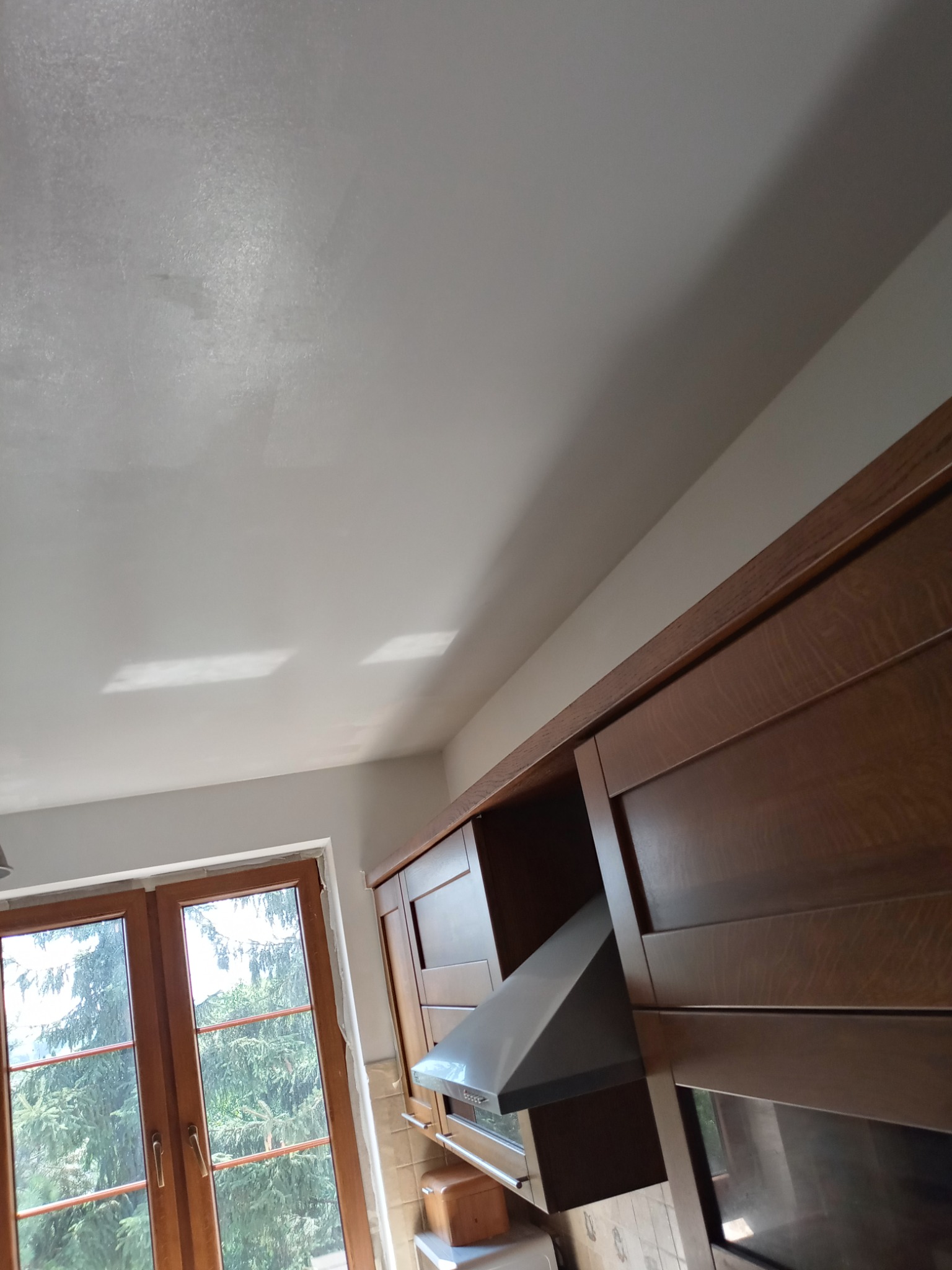 Wykończone wnętrze kuchni z widoczną gładzią na suficie, brązowe szafki kuchenne i okno z widokiem na zieleń.