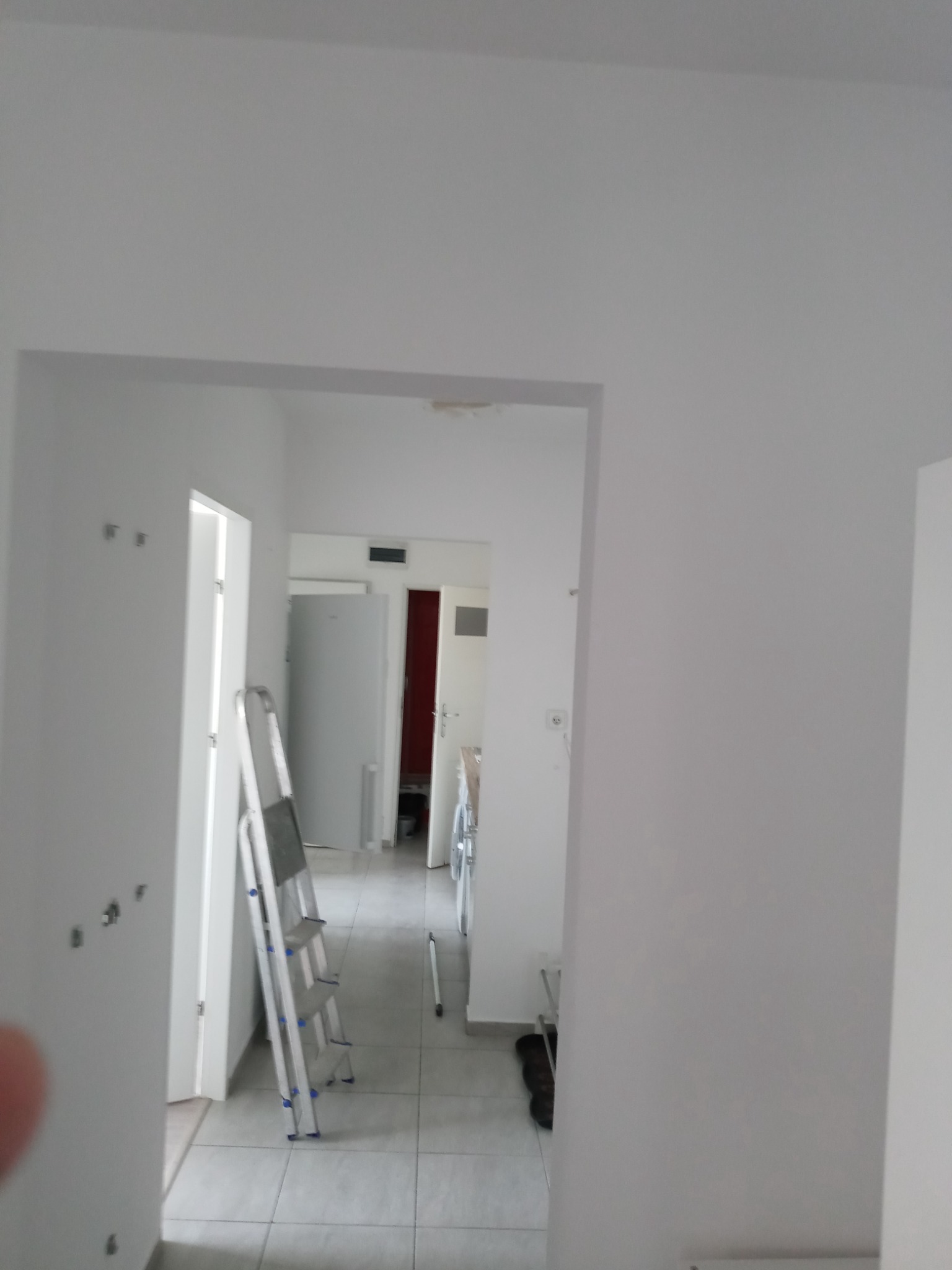 Wnętrze mieszkania w trakcie odświeżania, widoczne białe ściany, aluminiowa drabina oparta o framugę drzwi, szara podłoga z płytek, oraz widok na dalsze pomieszczenia.