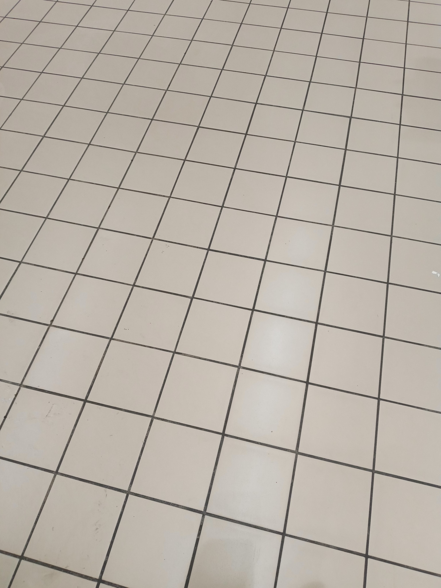 Beżowe, kwadratowe płytki podłogowe z ciemnoszarą fugą, ułożone ukośnie, widoczne drobne zabrudzenia.