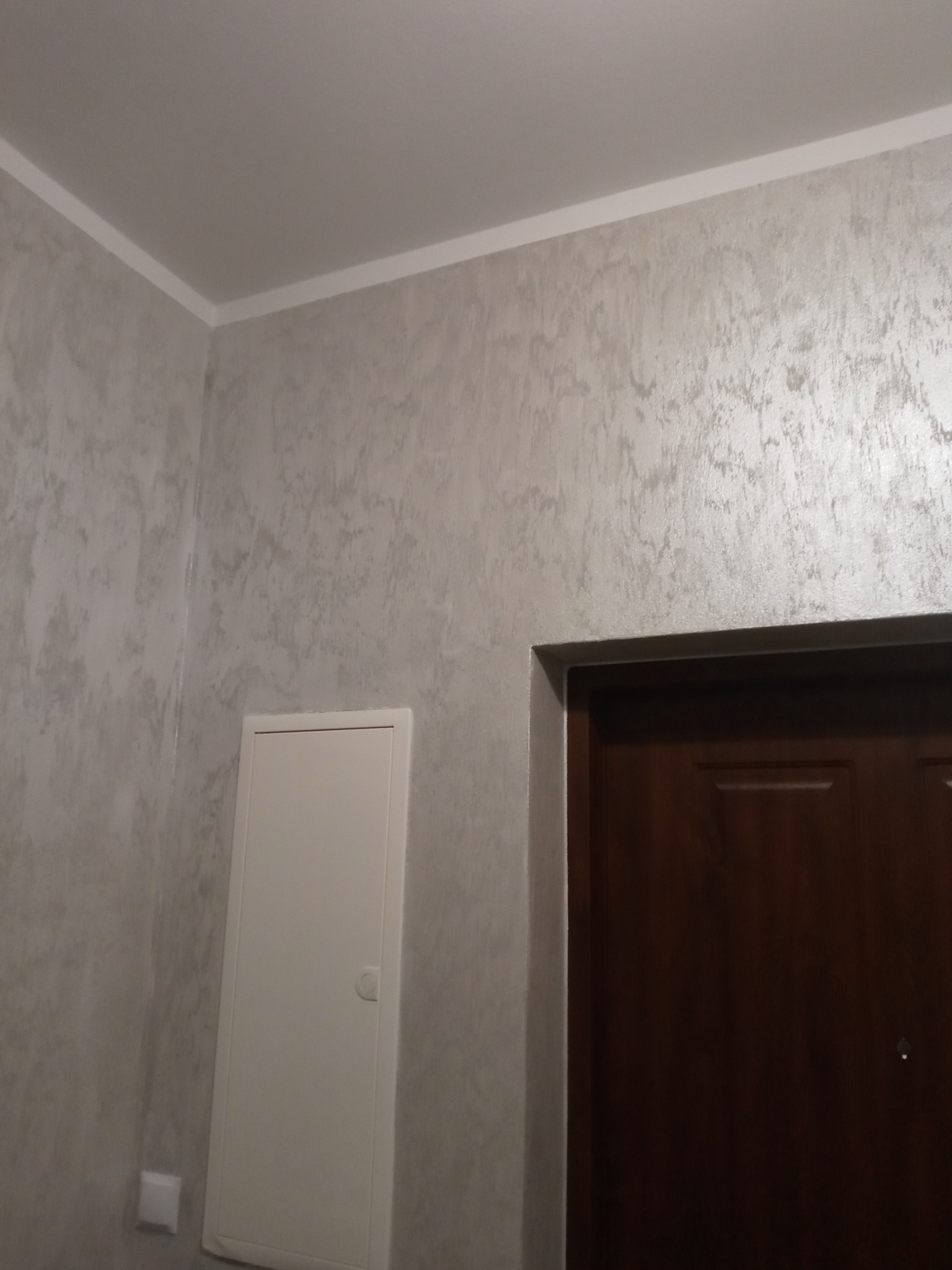 Ściana wewnętrzna wykończona dekoracyjną, srebrzystą fakturą, widoczny fragment brązowych drzwi i biała skrzynka rewizyjna.