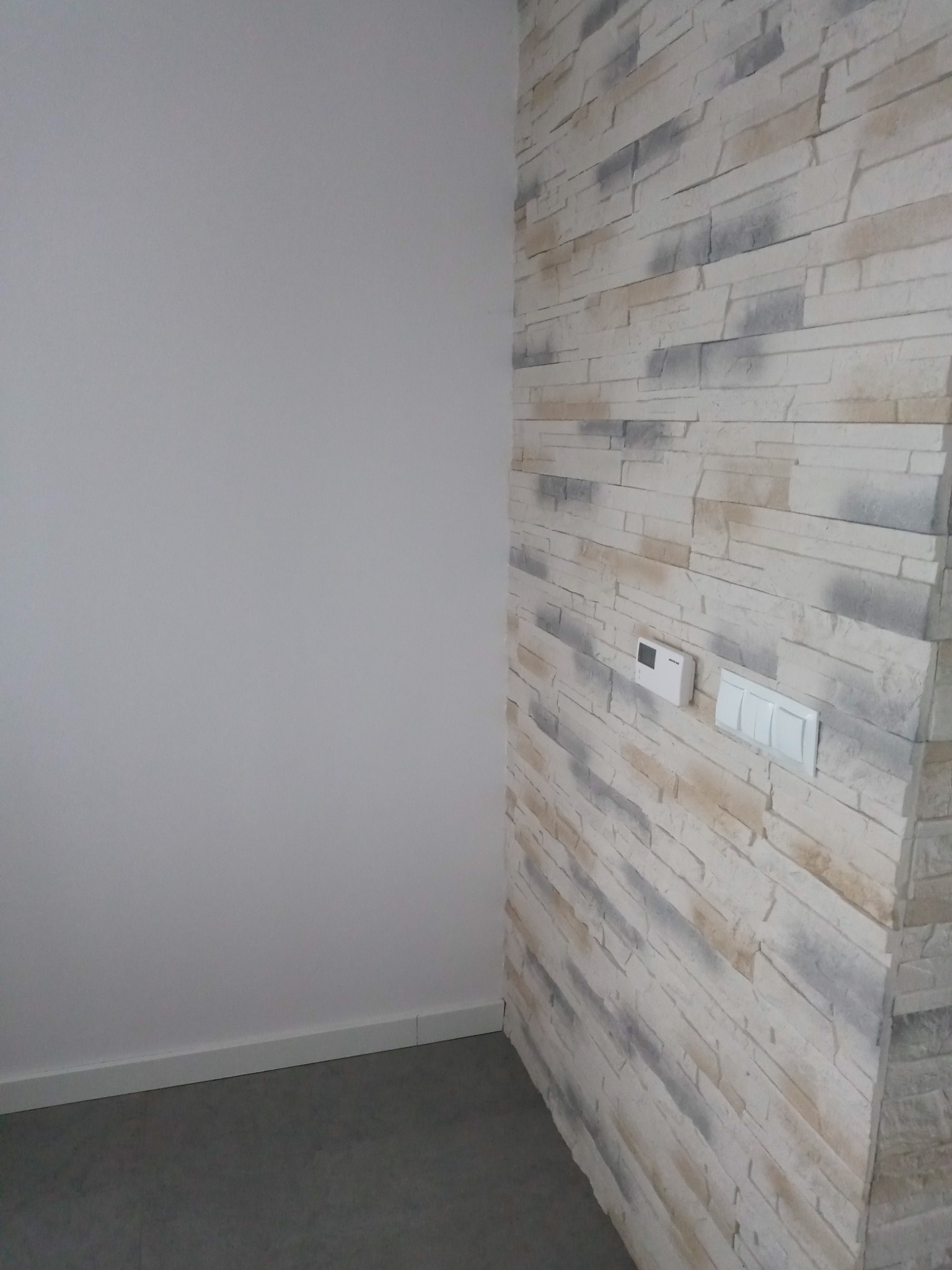 Ściana wewnętrzna wykończona dekoracyjną okładziną kamienną w odcieniach bieli, szarości i beżu, z widocznymi włącznikami światła i panelem sterowania klimatyzacją, sąsiadująca z gładką, pomalowaną...
