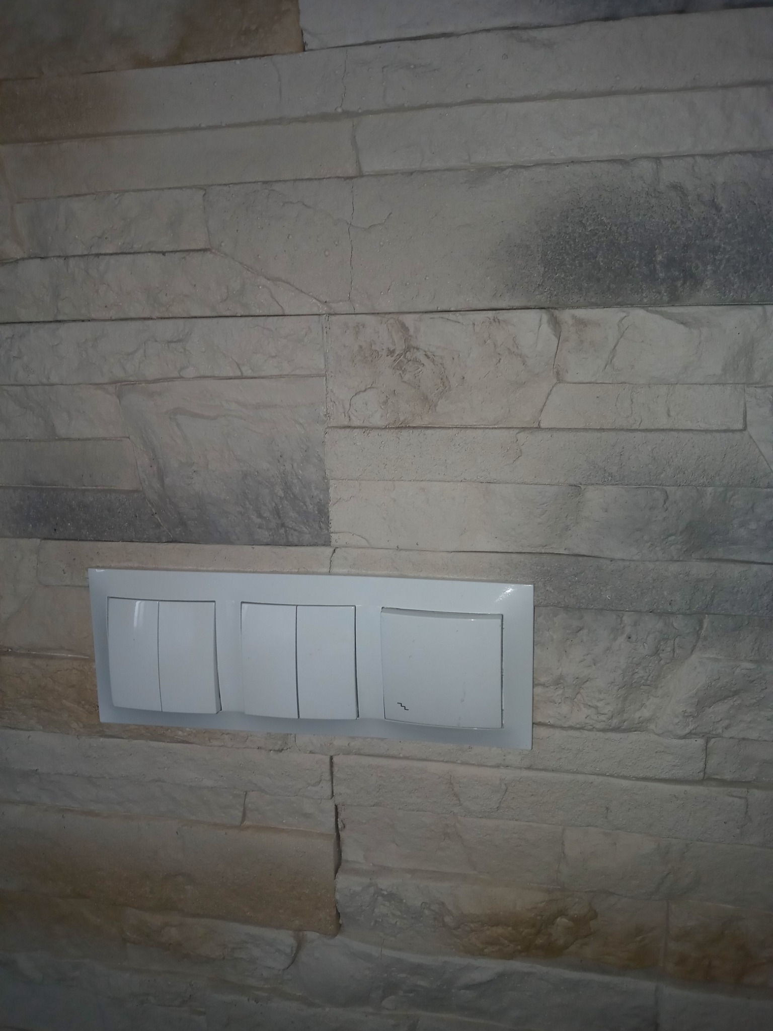 Ściana wykończona dekoracyjną okładziną imitującą kamień z zamontowanym białym, potrójnym włącznikiem światła.