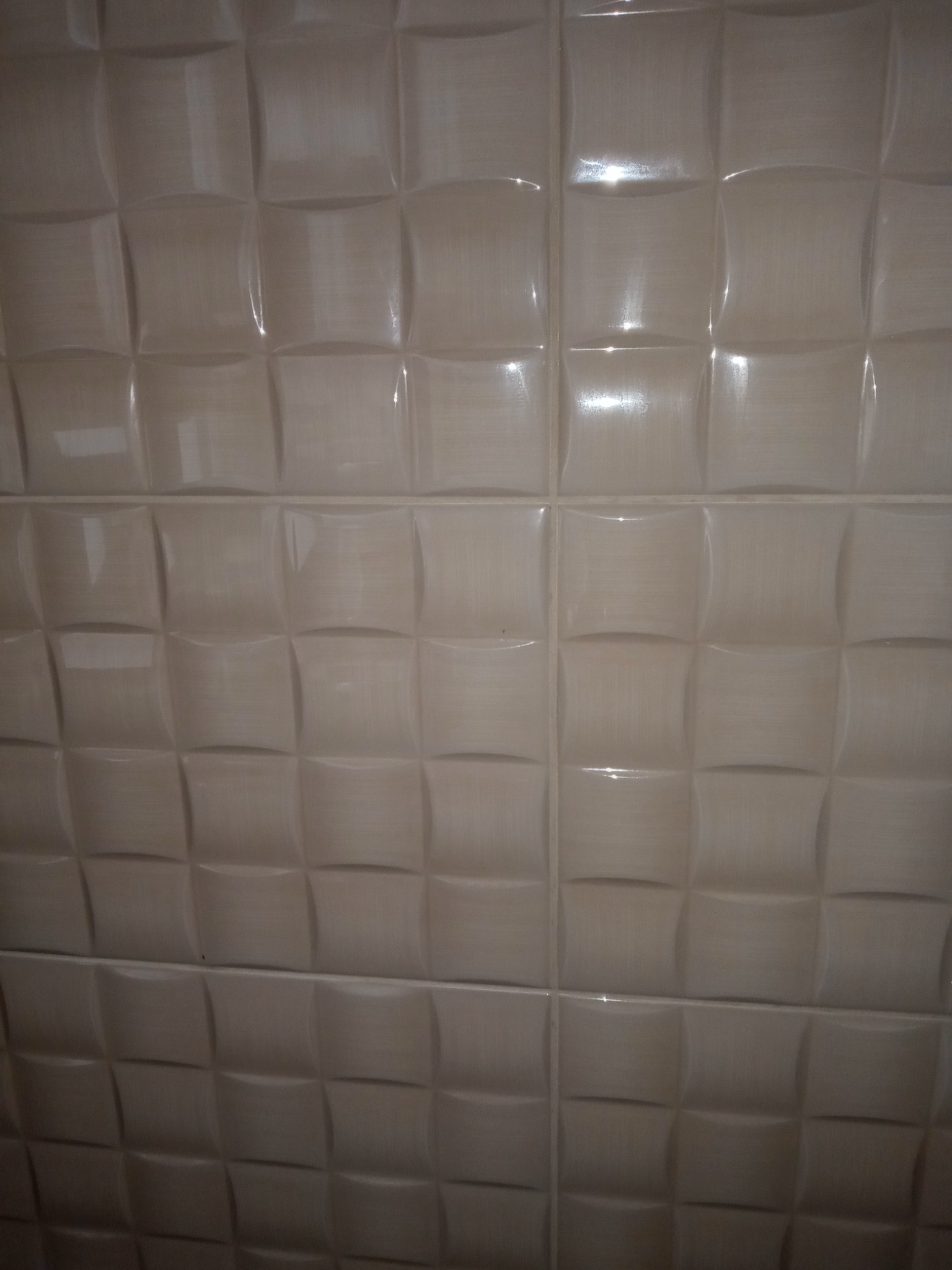 Ściana wyłożona beżowymi, wypukłymi płytkami ceramicznymi z błyszczącą powierzchnią, ułożonymi równo w kwadratową siatkę.