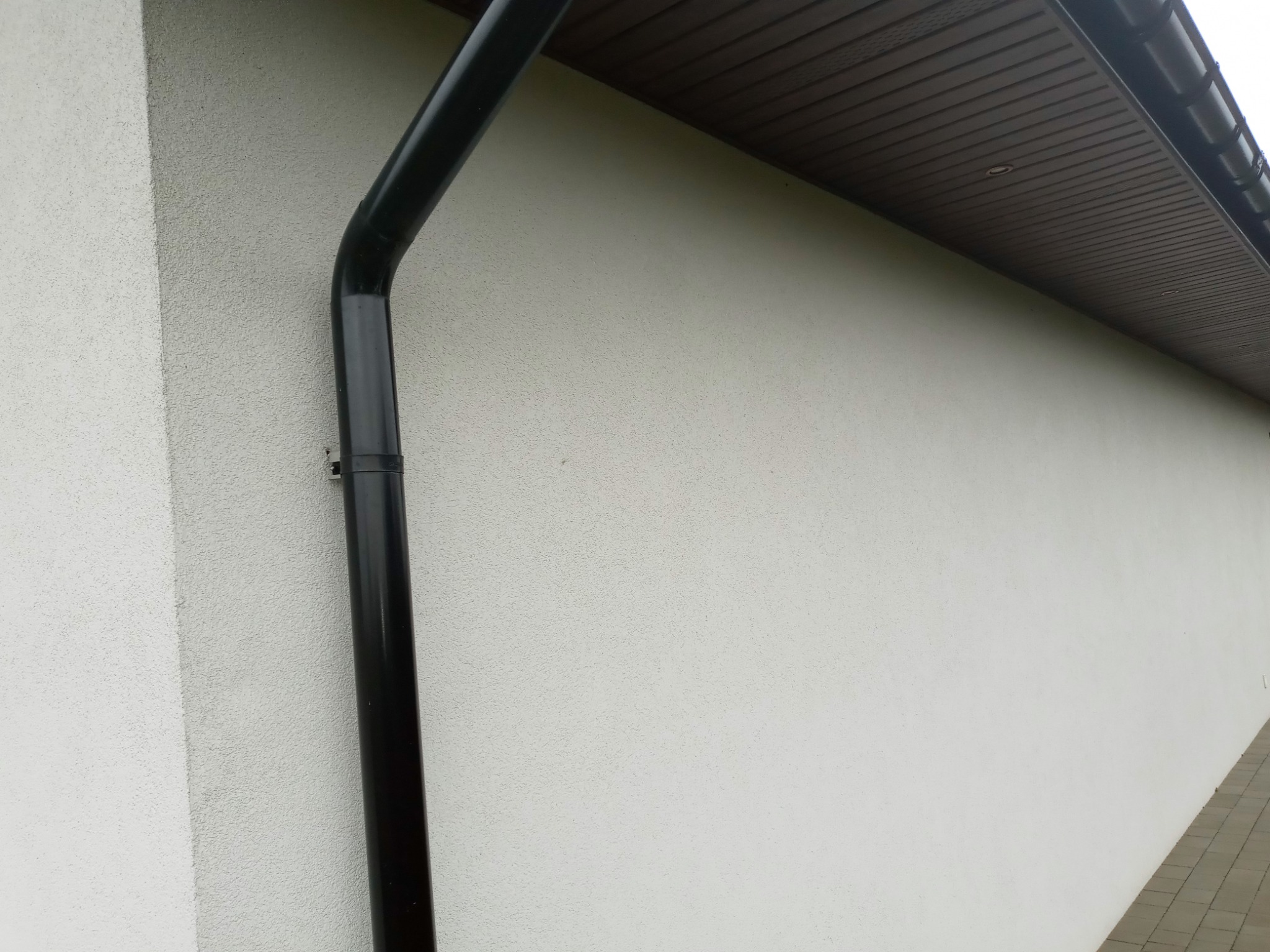 Fragment jasnej elewacji budynku z widoczną czarną rynną spustową i brązowym podbitką dachową.