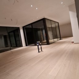 SPRINTWOOD SPÓŁKA Z OGRANICZONĄ ODPOWIEDZIALNOŚCIĄ - Jasna drewniana podłoga w przestronnym wnętrzu z dużymi oknami, widoczne niedokończone prace wykończeniowe, wystające kable z sufitu i narzędzia na podłodze.