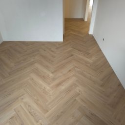 SPRINTWOOD SPÓŁKA Z OGRANICZONĄ ODPOWIEDZIALNOŚCIĄ - Jasne panele ułożone w jodełkę w pustym, świeżo wyremontowanym pokoju z białymi ścianami. Widoczny fragment gniazdka elektrycznego.