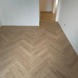 Jasne panele ułożone w jodełkę w pustym, świeżo wyremontowanym pokoju z białymi ścianami. Widoczny fragment gniazdka elektrycznego.