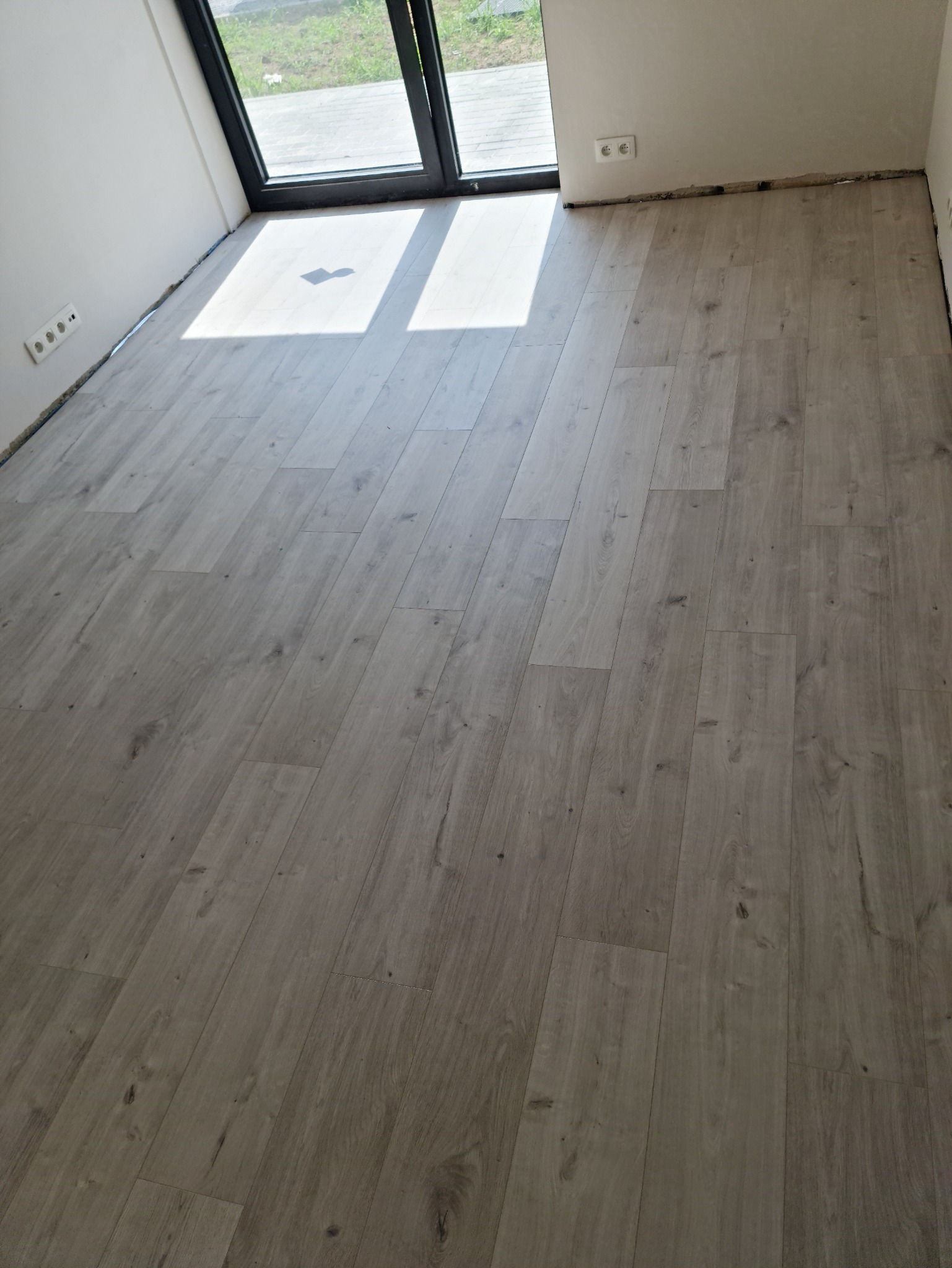 Jasnoszara podłoga z paneli ułożona w nowym pomieszczeniu, widoczne gniazdka elektryczne i fragment okna balkonowego z widokiem na zewnątrz.