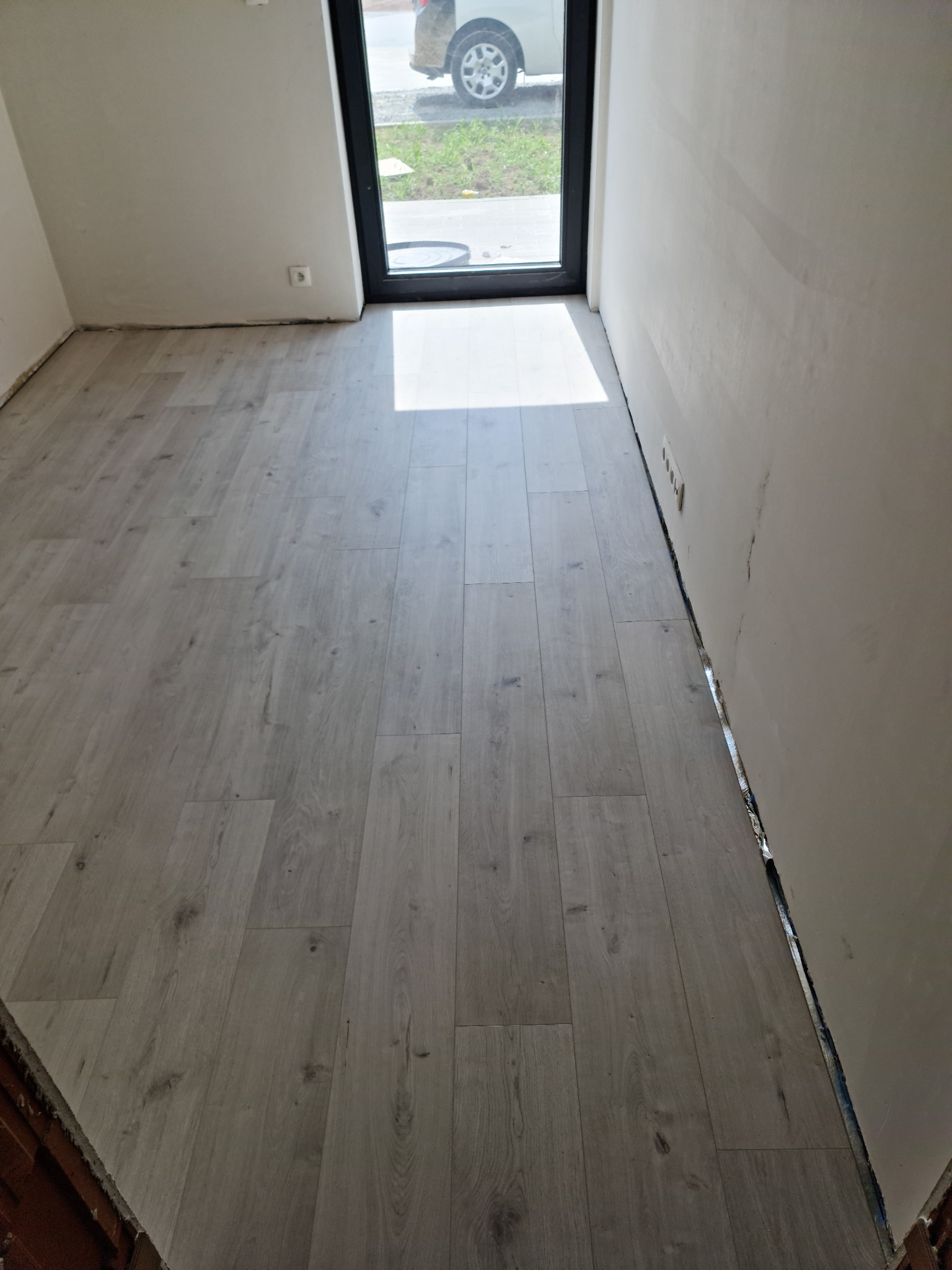 Świeżo ułożone, jasne panele podłogowe w pomieszczeniu w Nieszawie, widok na okno z fragmentem samochodu na zewnątrz, ściany przygotowane do malowania.