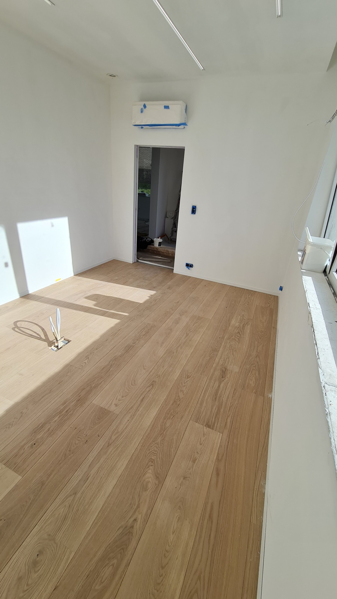 Jasne panele podłogowe ułożone w pustym, białym pomieszczeniu z wystającymi przewodami w podłodze i klimatyzatorem zabezpieczonym folią malarską.