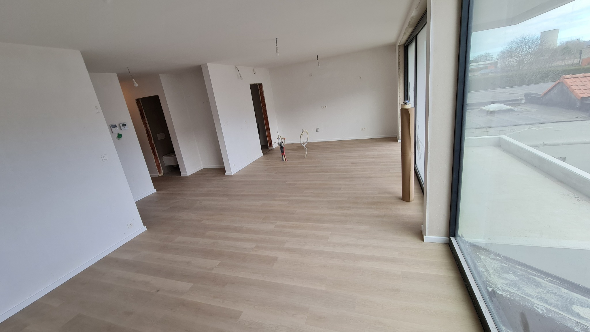 Jasne panele podłogowe ułożone w pustym, jasnym pomieszczeniu z dużym oknem, widoczne wystające przewody elektryczne i narzędzia do montażu listew przypodłogowych.