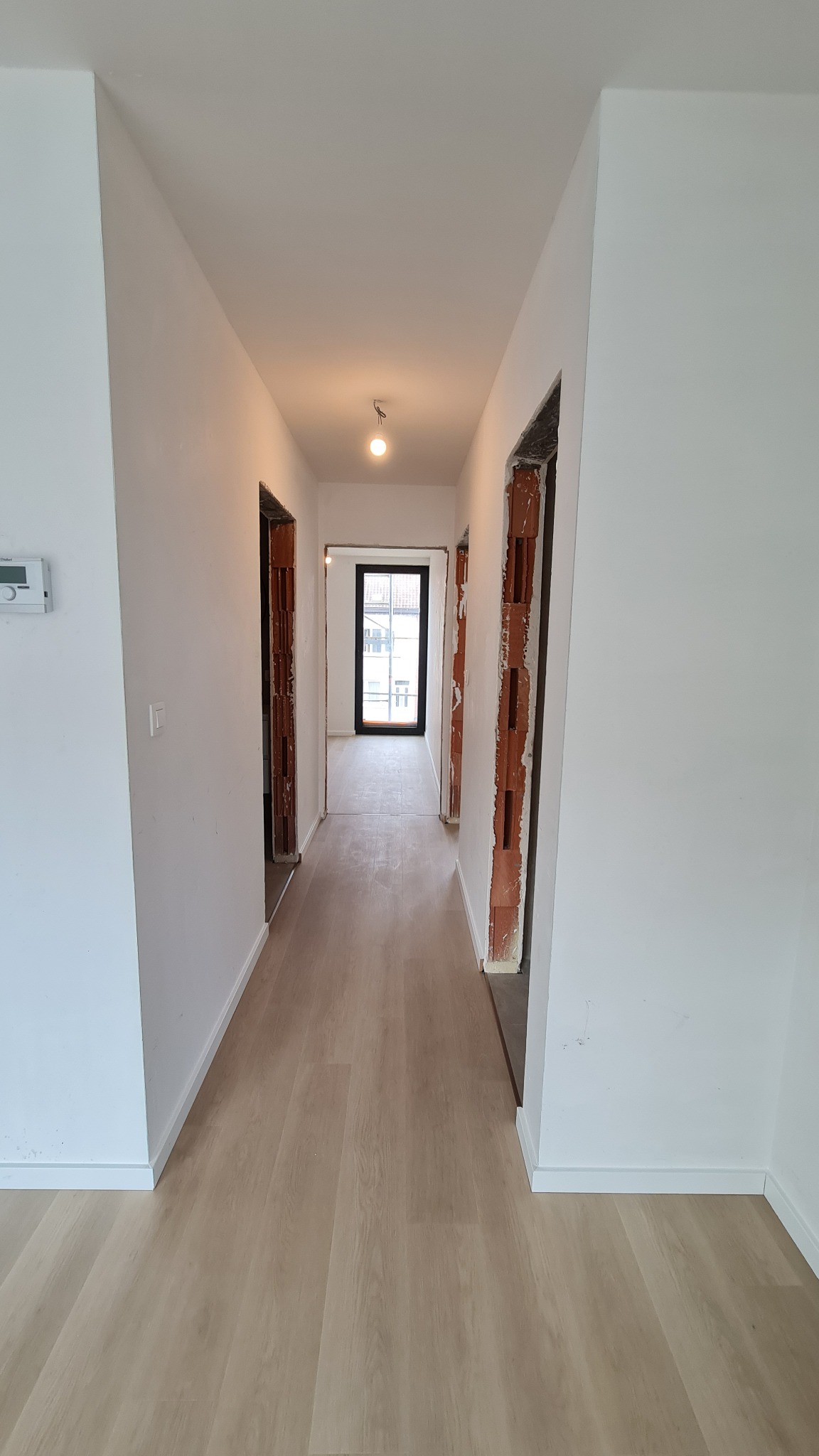 Korytarz w trakcie remontu z jasnymi panelami podłogowymi, widoczne surowe framugi drzwiowe z ceglanymi wstawkami i białe ściany. Na ścianie widoczny regulator temperatury.