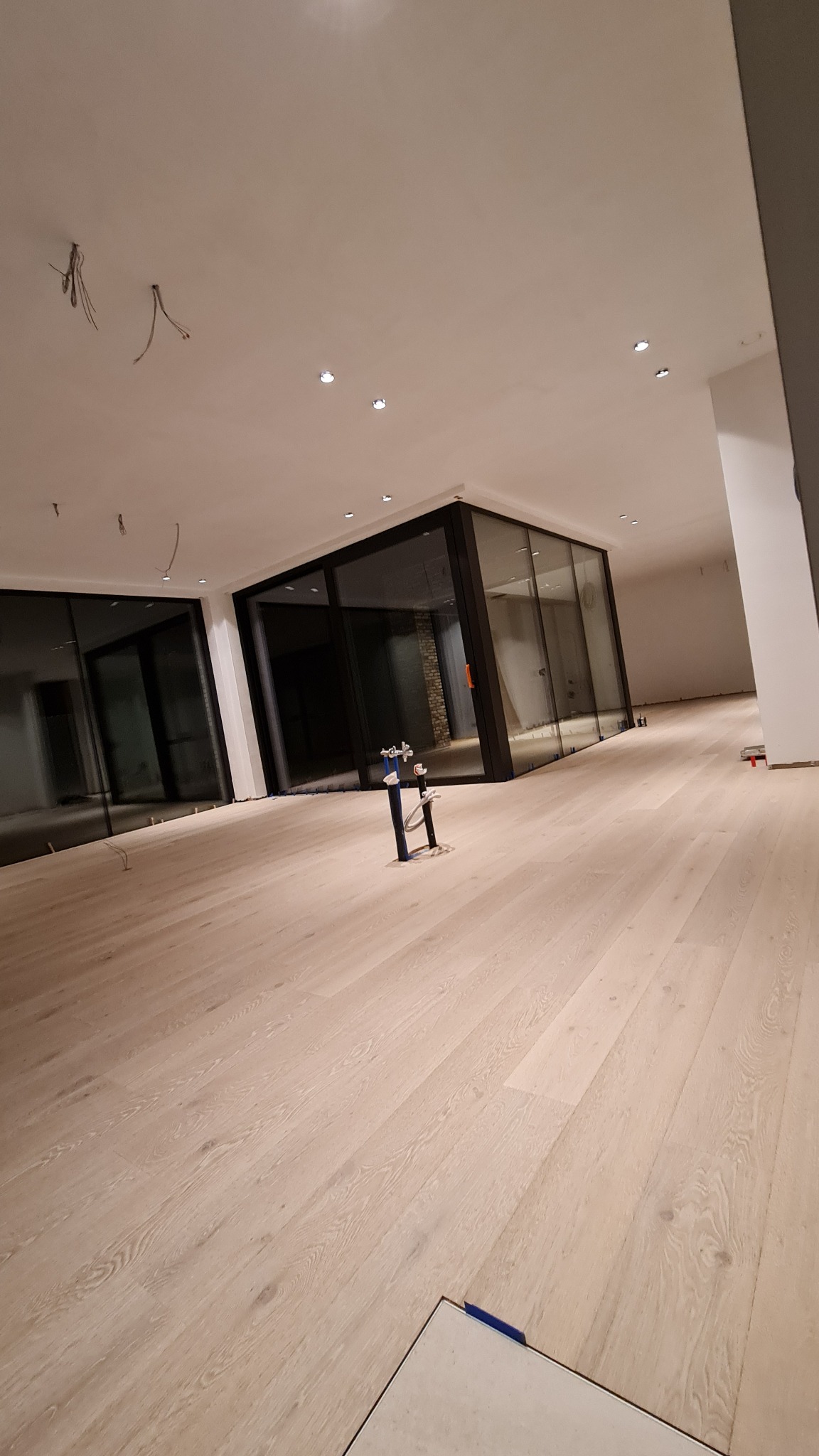 Jasna drewniana podłoga w przestronnym wnętrzu z dużymi oknami, widoczne niedokończone prace wykończeniowe, wystające kable z sufitu i narzędzia na podłodze.