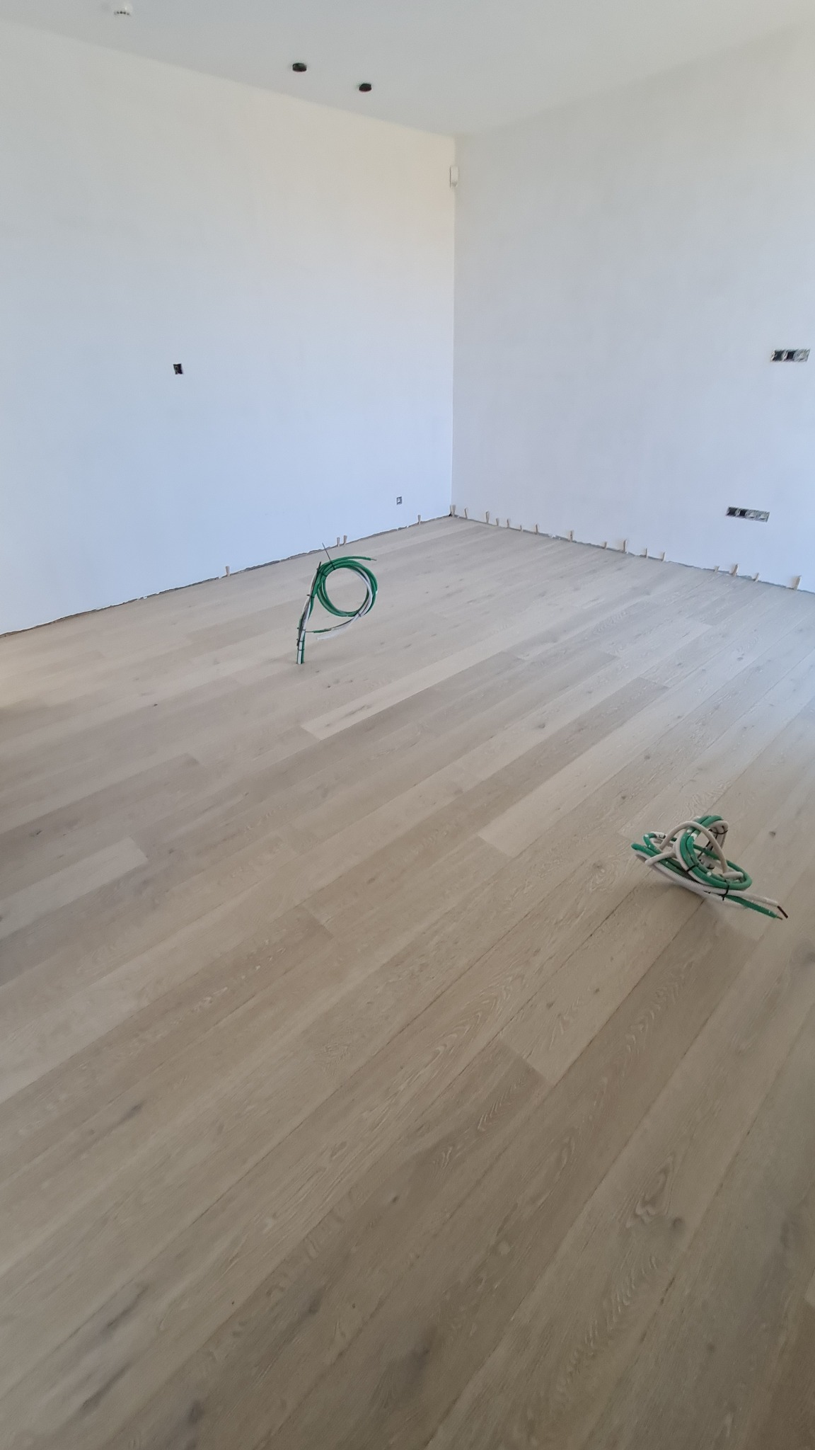 Jasny parkiet ułożony w nowym pomieszczeniu, widoczne kable elektryczne wystające z podłogi i listwy przypodłogowe w trakcie montażu.